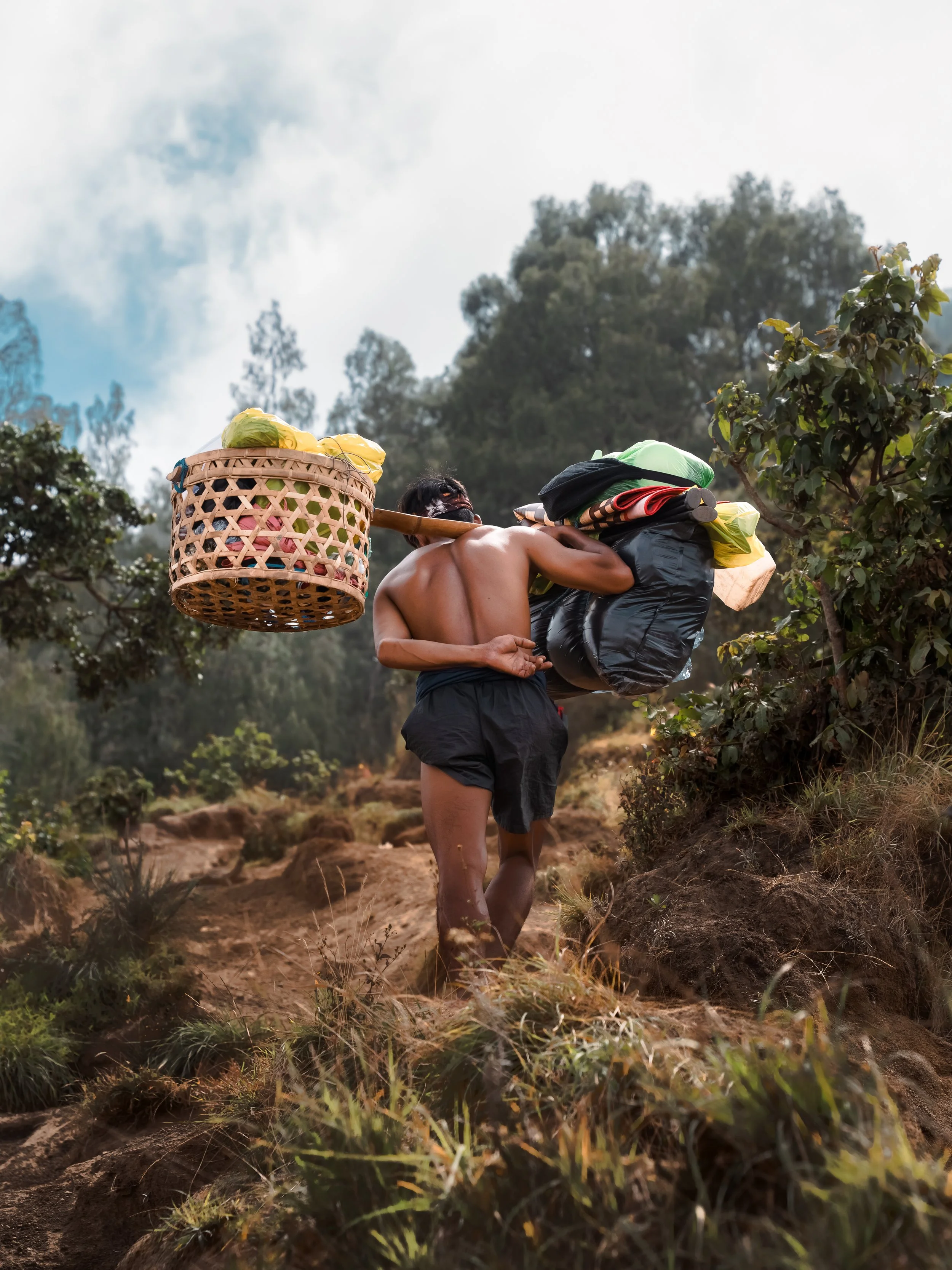 Een persoon met een rugzak en een mand op de schouders wandelt door een heuvelachtig landschap met bomen en struiken.