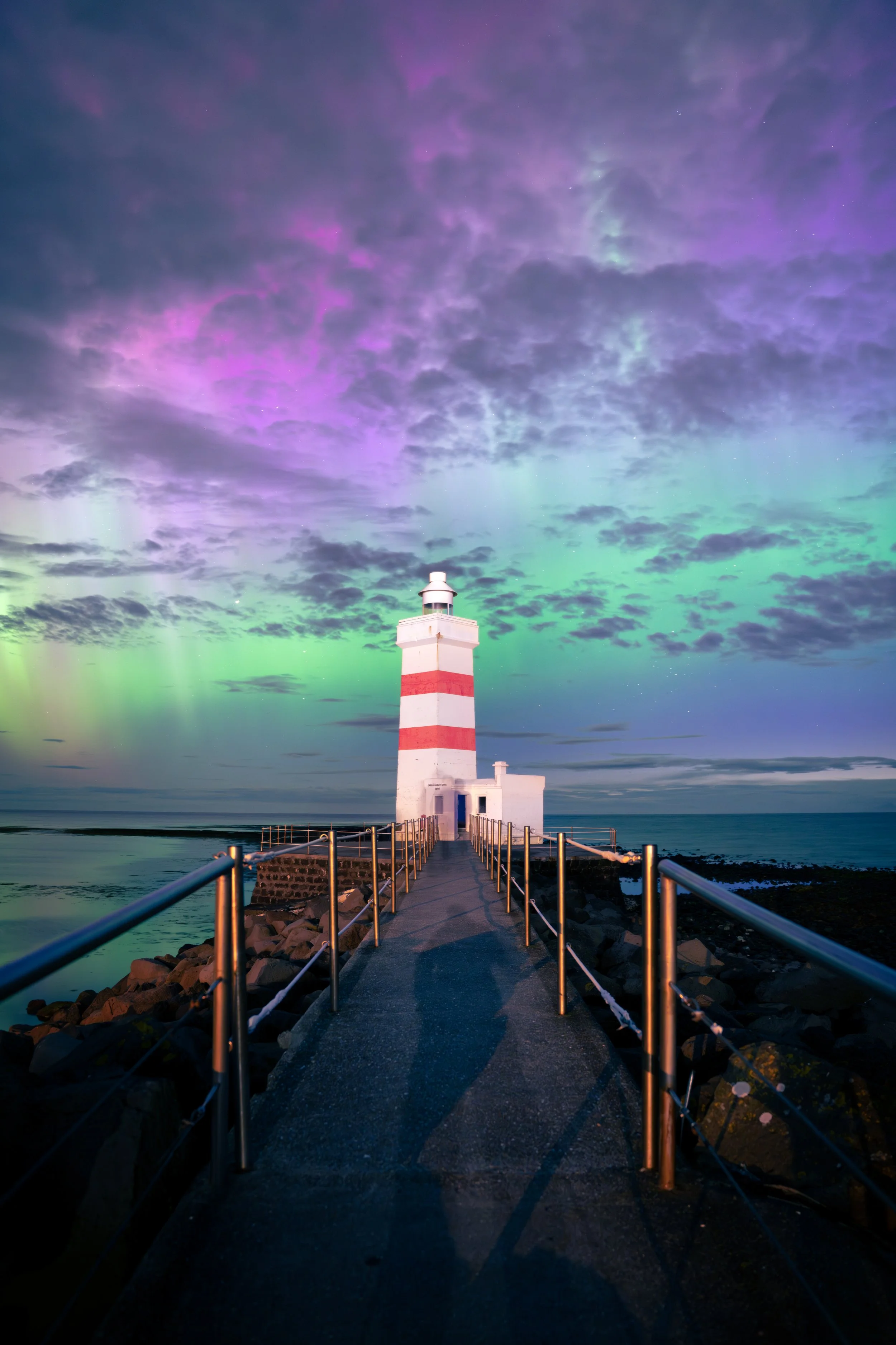 Een vuurtoren met rood en wit horizontale strepen staat op een rotseiland, met een pad en hek eromheen, onder een kleurrijke hemel met noorderlicht en wolken.