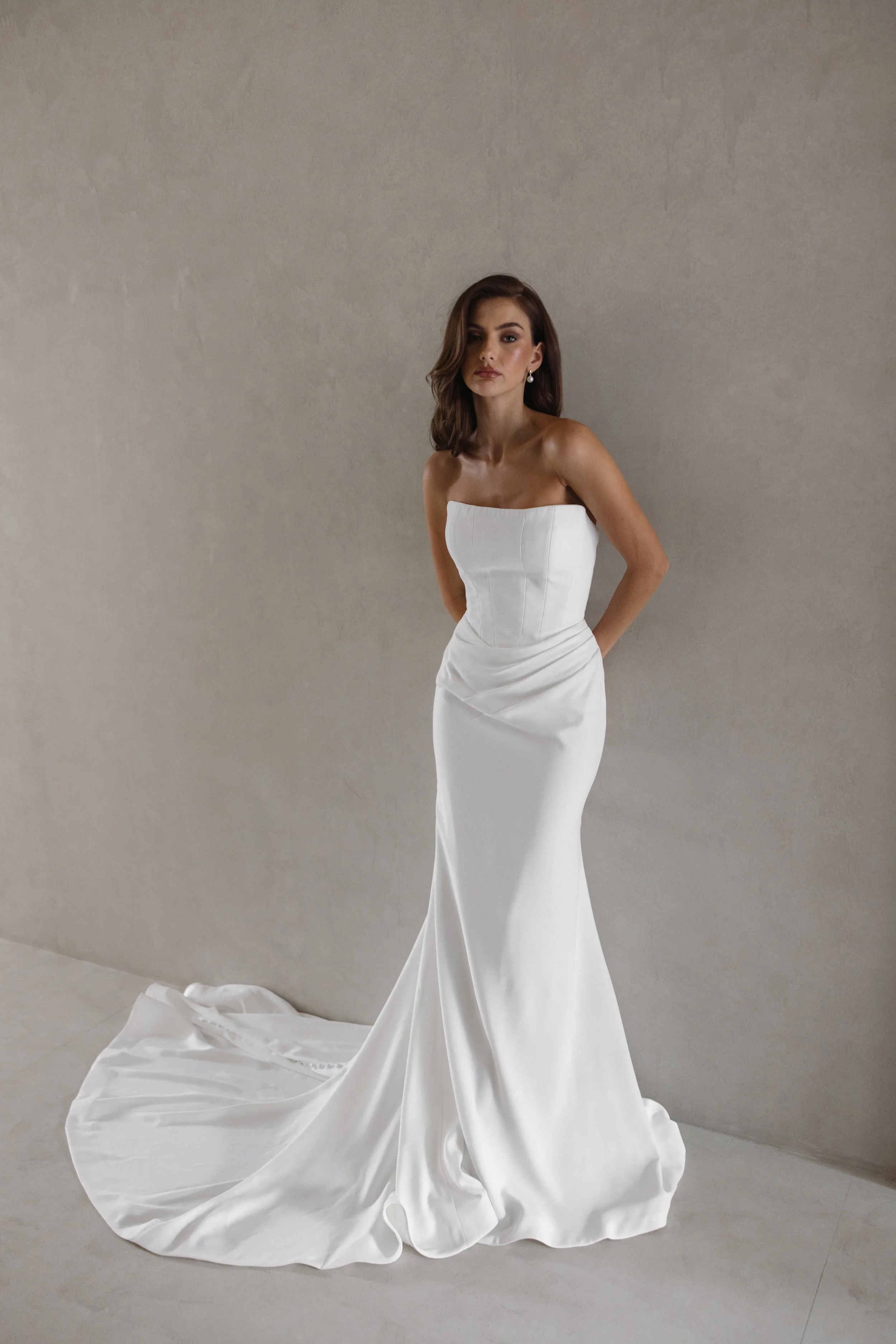A woman in a strapless white wedding dress with a train stands against a neutral wall.