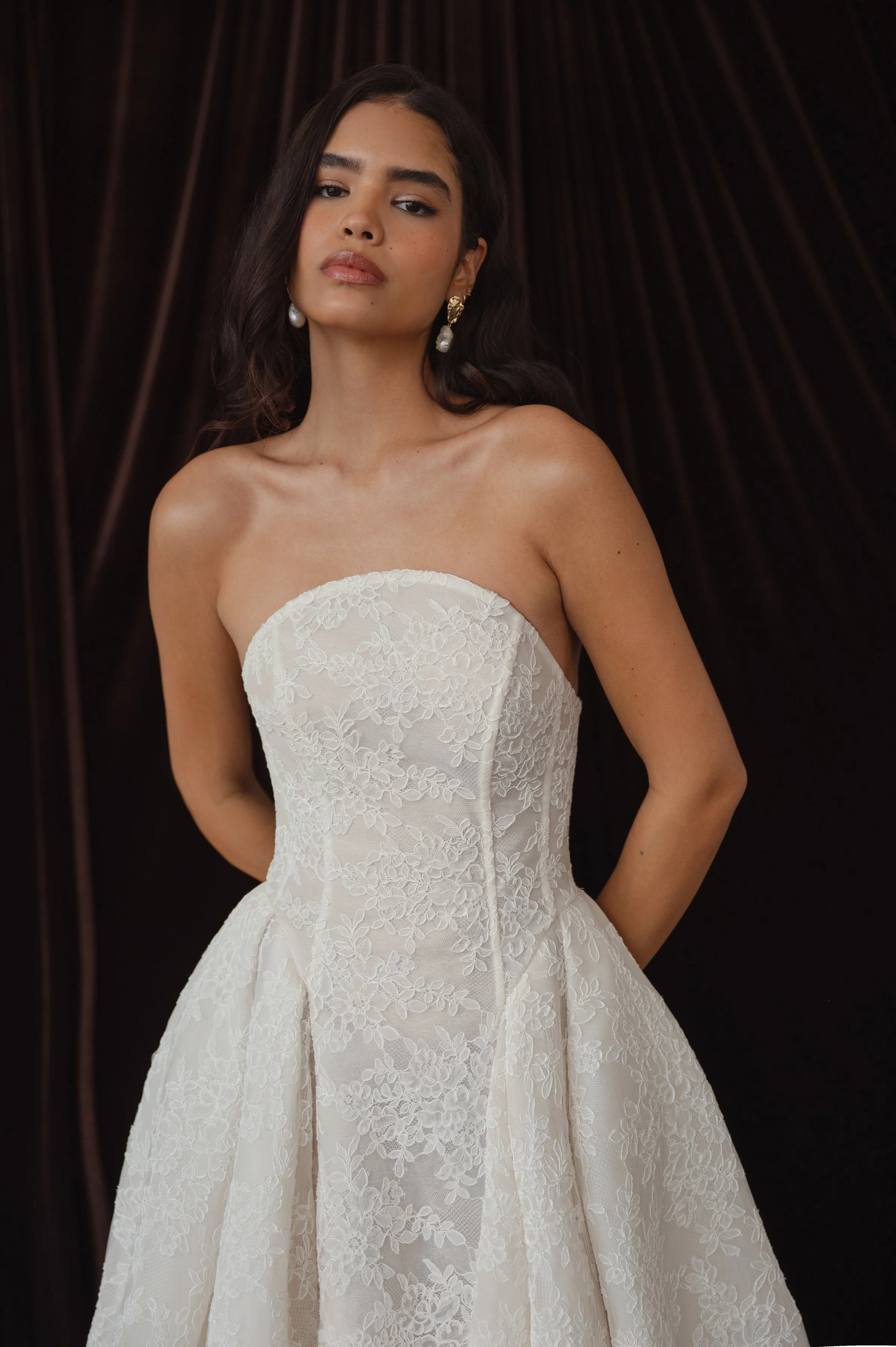 The Jenny Yoo Ember gown is a dramatic lace ball gown cut in intricately textured Chappell lace with a modern arched neckline and subtle drop-waist seaming that balances romantic tradition with contemporary flair. It features a full, pleated skirt wi
