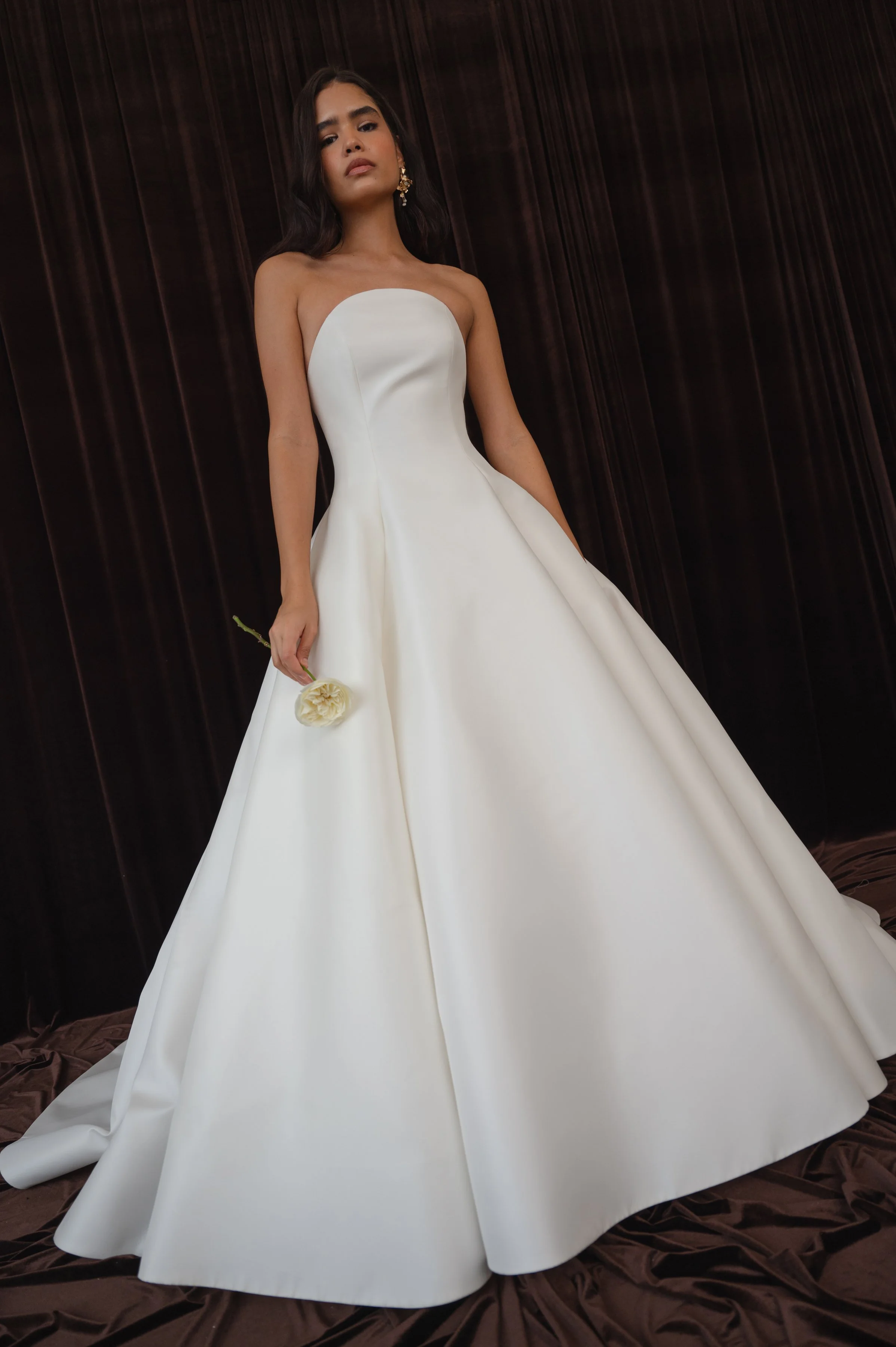 The Jenny Yoo Bridal Maya gown with applique is a modern yet timeless ball gown with a striking arched neckline and clean, elegant lines, anchored by a dramatic full skirt. Bold, oversized floral appliques artfully adorn the hem of the skirt, giving 