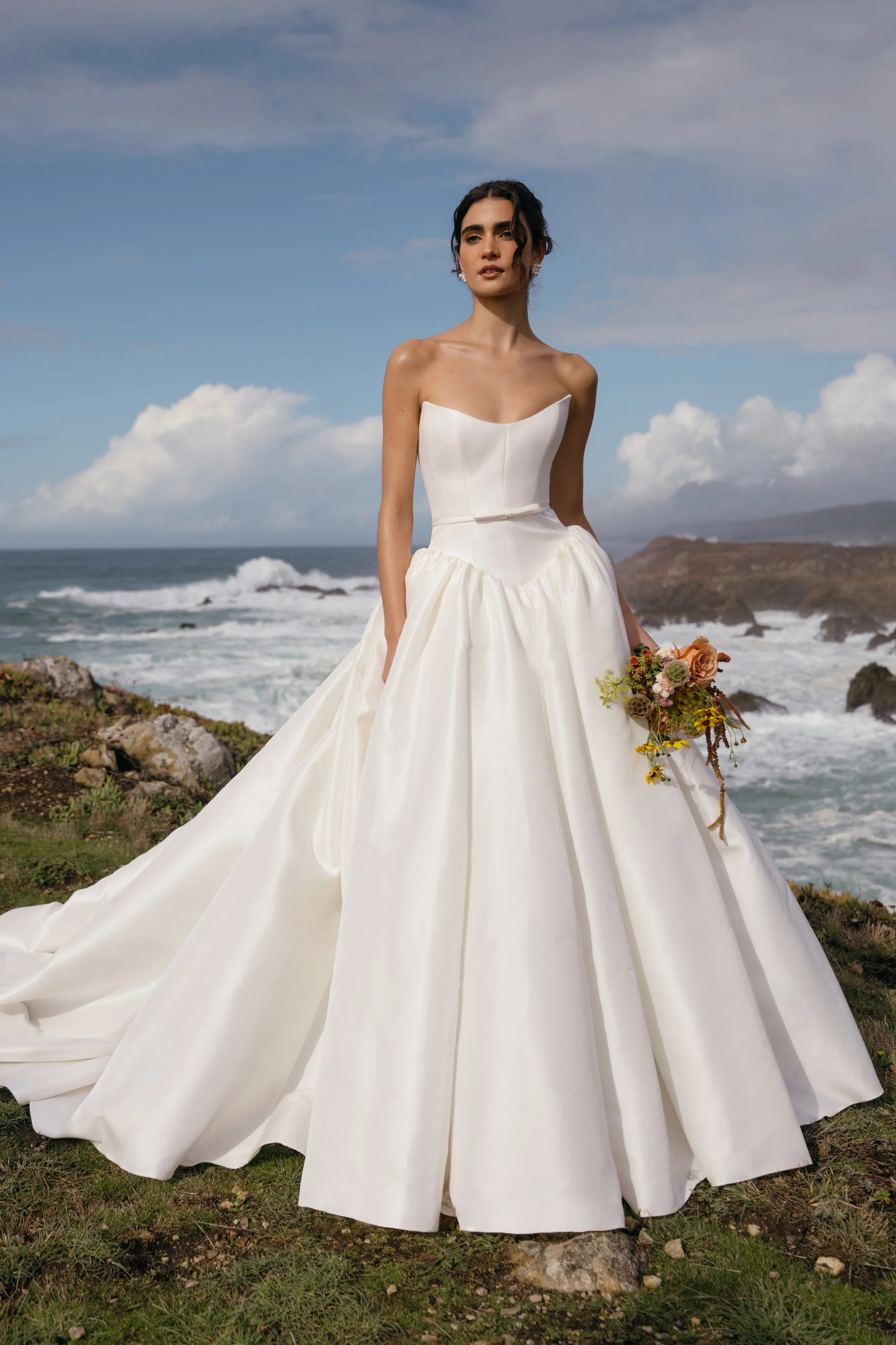 The Jenny Yoo Danielle gown is a romantic Italian taffeta ball gown with a fully corseted bodice and a striking high, peaked scoop neckline that creates a confident, flattering silhouette. It’s balanced by a voluminous gathered skirt, delicate bow sa
