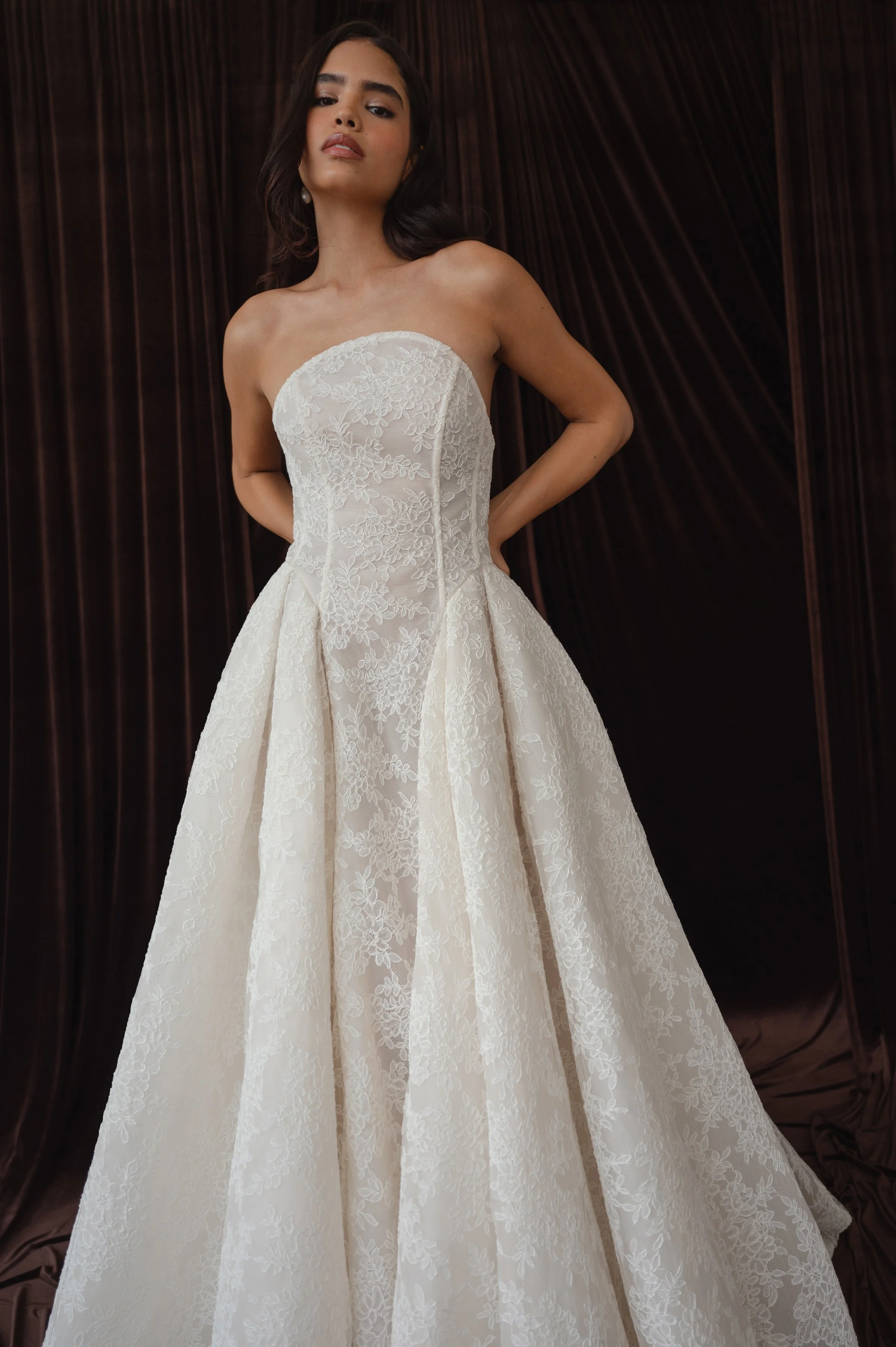 The Jenny Yoo Ember gown is a dramatic lace ball gown cut in intricately textured Chappell lace with a modern arched neckline and subtle drop-waist seaming that balances romantic tradition with contemporary flair. It features a full, pleated skirt wi