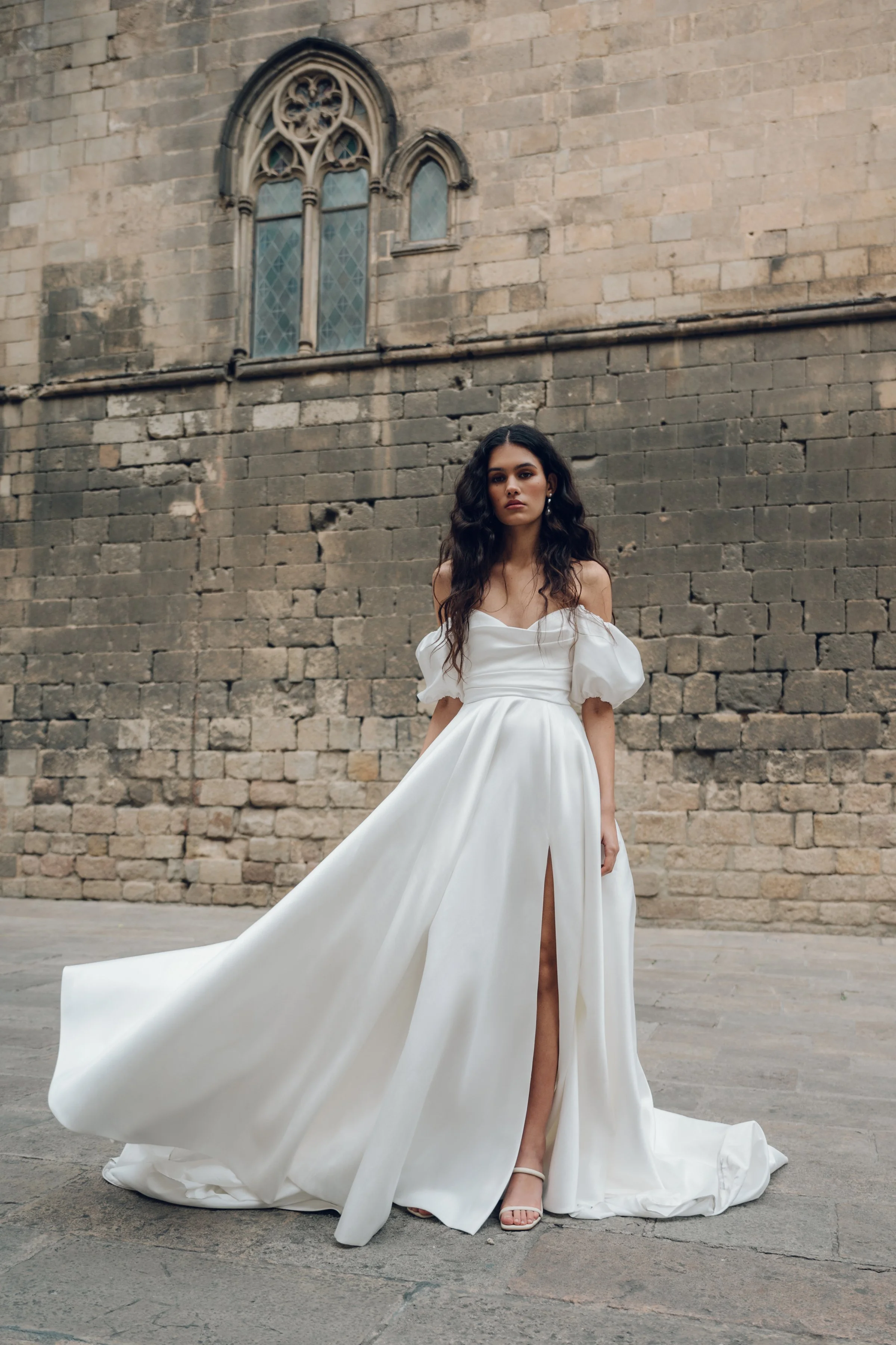 The Jenny Yoo Marisol gown is a romantic satin ball gown with a beautifully draped bodice and a sultry, sculpted cowl neckline that feels effortlessly feminine. It features a full skirt interrupted by a flattering slit and comes with detachable puff 