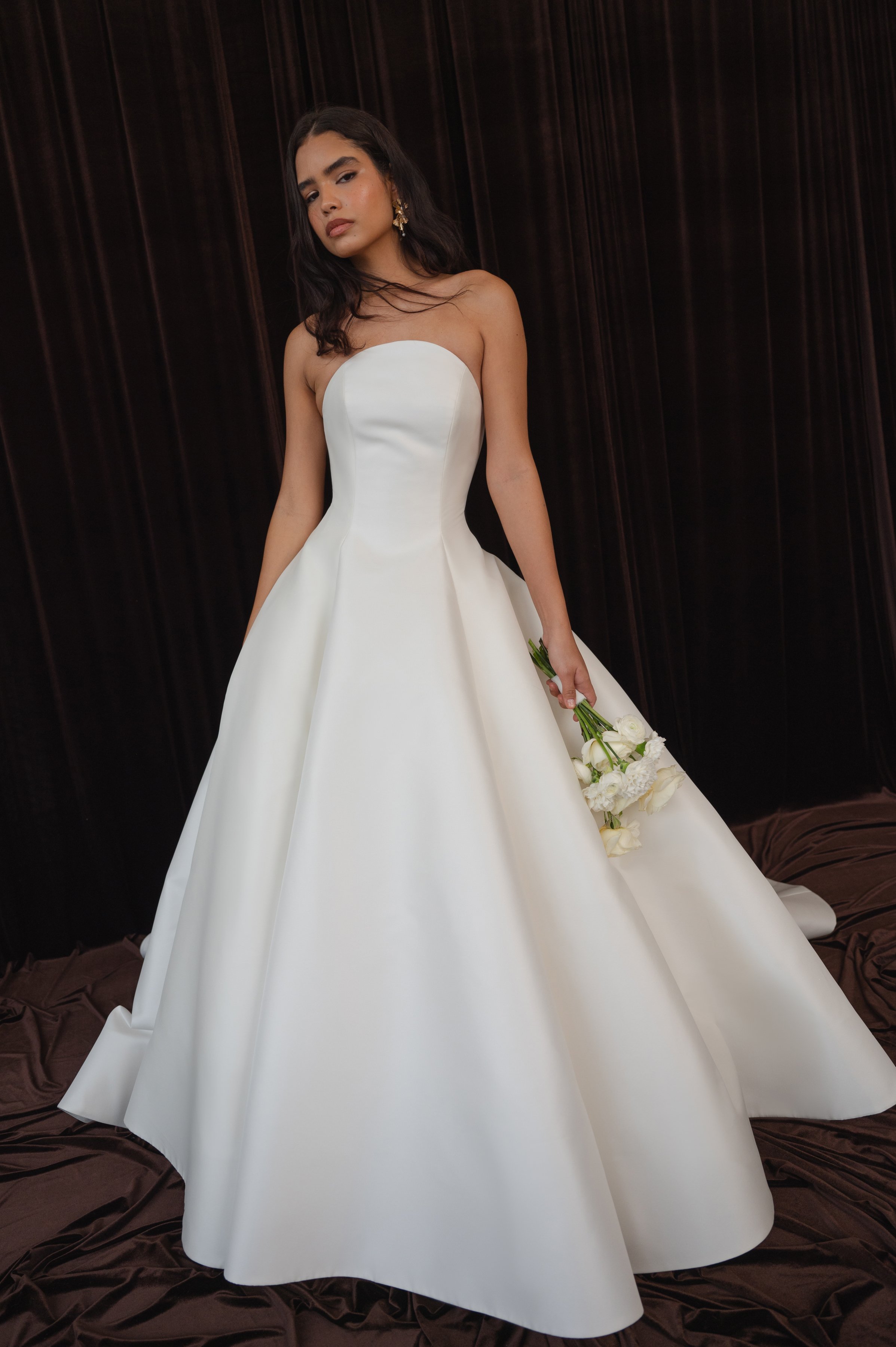 The Jenny Yoo Bridal Maya gown with applique is a modern yet timeless ball gown with a striking arched neckline and clean, elegant lines, anchored by a dramatic full skirt. Bold, oversized floral appliques artfully adorn the hem of the skirt, giving 