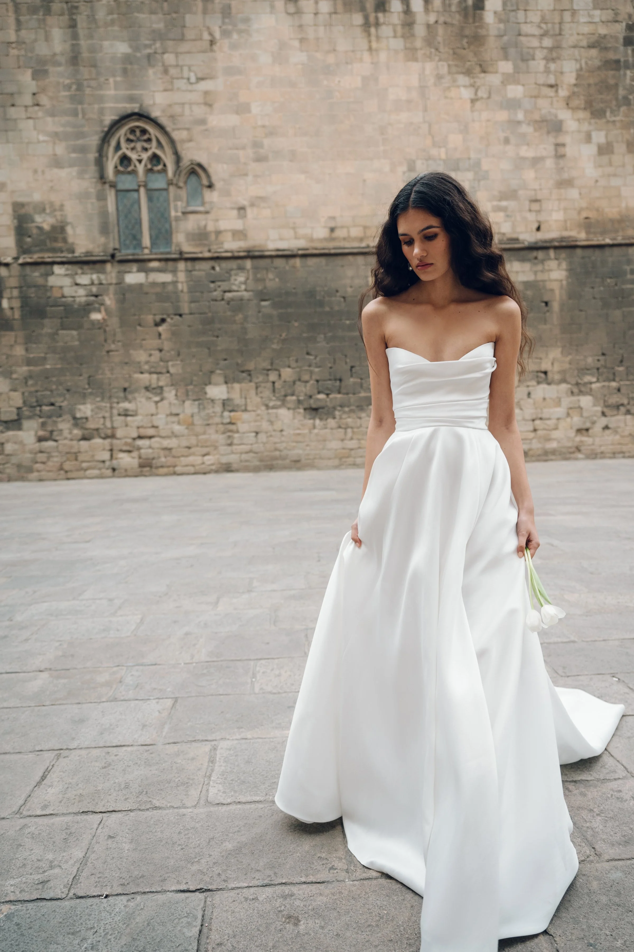 The Jenny Yoo Marisol gown is a romantic satin ball gown with a beautifully draped bodice and a sultry, sculpted cowl neckline that feels effortlessly feminine. It features a full skirt interrupted by a flattering slit and comes with detachable puff 