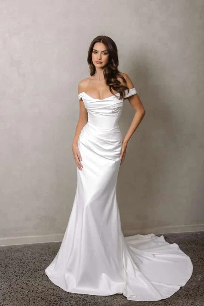 A woman in a white wedding gown with off-the-shoulder sleeves, standing against a plain wall.