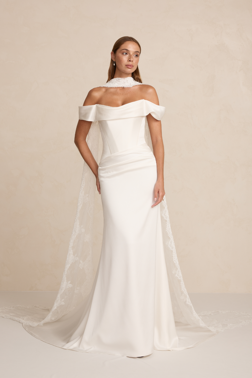 The MWL Brooks is crafted from luxurious satin crepe, featuring an asymmetrical waistline, a structured boned bodice, and a soft front cowl. A sleek column skirt with four pleats creates an elegant, elongated silhouette. Available at Halo and Wren Br