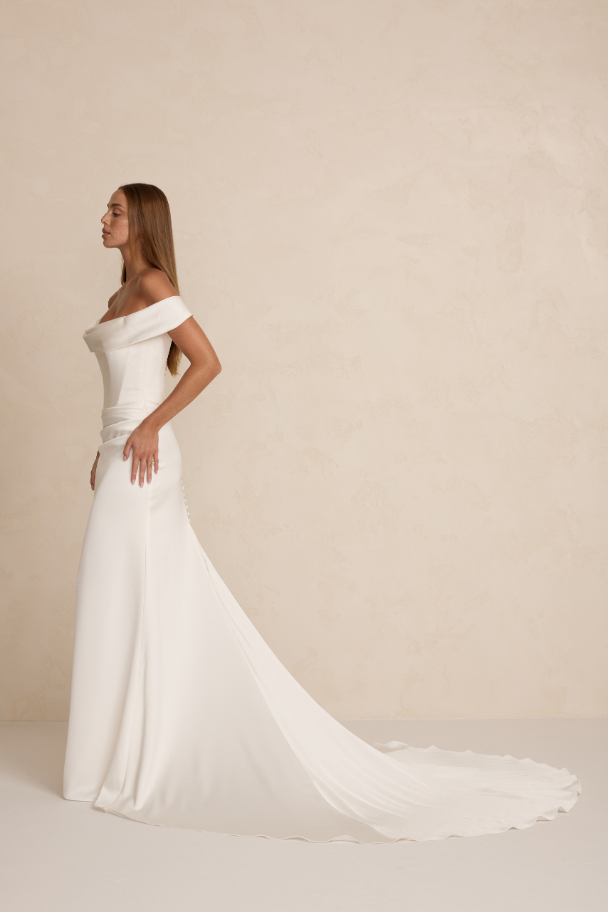 The MWL Brooks is crafted from luxurious satin crepe, featuring an asymmetrical waistline, a structured boned bodice, and a soft front cowl. A sleek column skirt with four pleats creates an elegant, elongated silhouette. Available at Halo and Wren Br