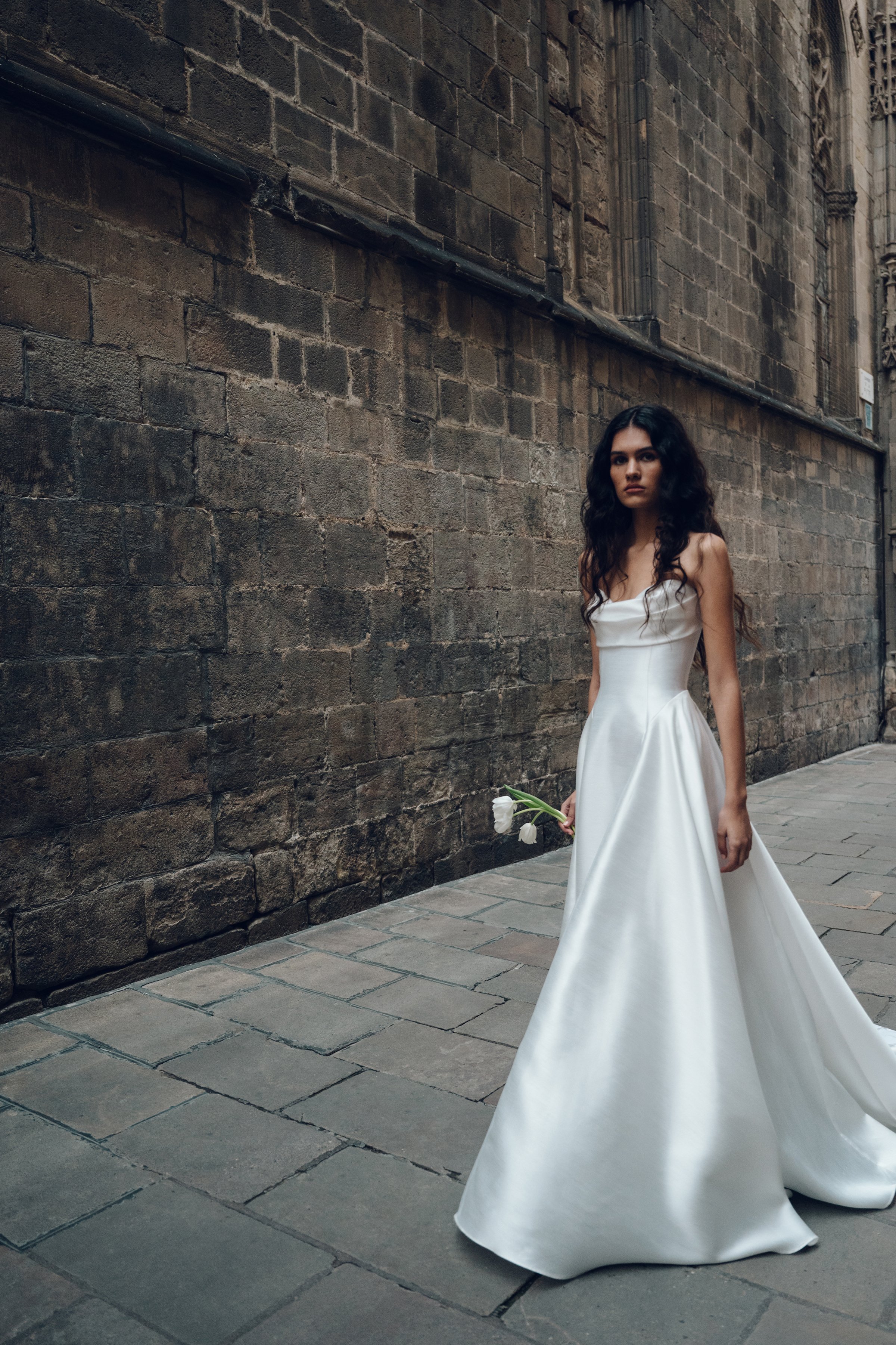 The Jenny Yoo Romina gown is a refined strapless A-line wedding dress crafted from lightweight, luminous canopy taffeta with a beautifully draped cowl bodice and sculpted drop-waist silhouette that feels both modern and timeless. Its soft, elegant li
