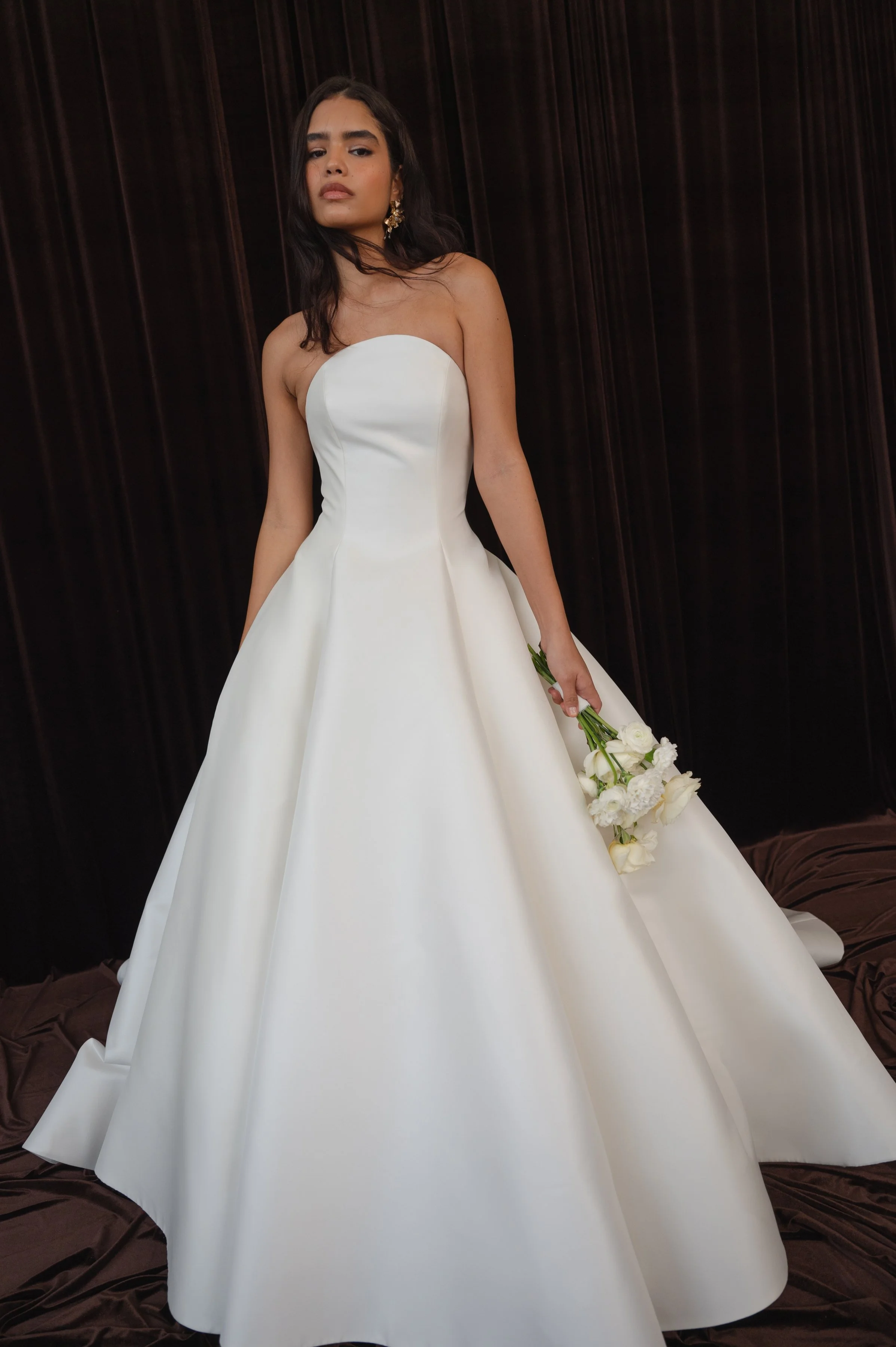 The Jenny Yoo Bridal Maya gown with applique is a modern yet timeless ball gown with a striking arched neckline and clean, elegant lines, anchored by a dramatic full skirt. Bold, oversized floral appliques artfully adorn the hem of the skirt, giving 