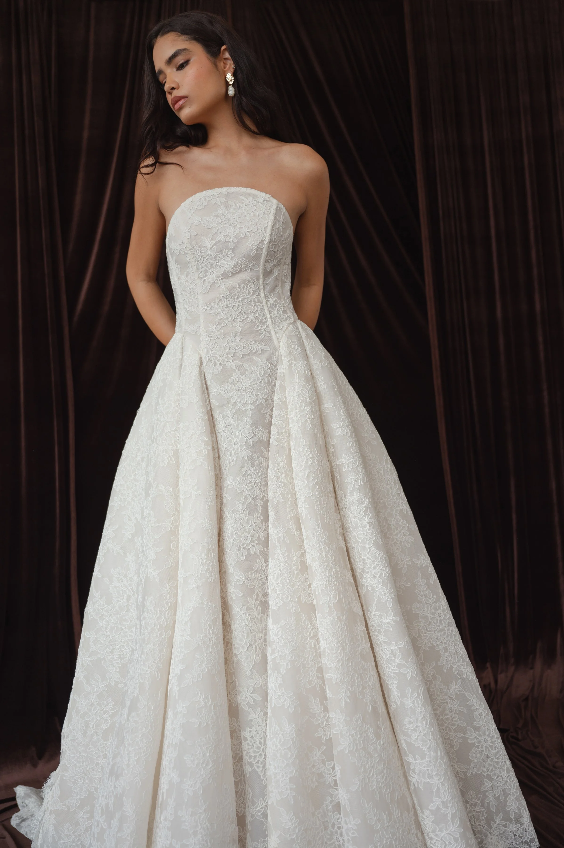 The Jenny Yoo Ember gown is a dramatic lace ball gown cut in intricately textured Chappell lace with a modern arched neckline and subtle drop-waist seaming that balances romantic tradition with contemporary flair. It features a full, pleated skirt wi