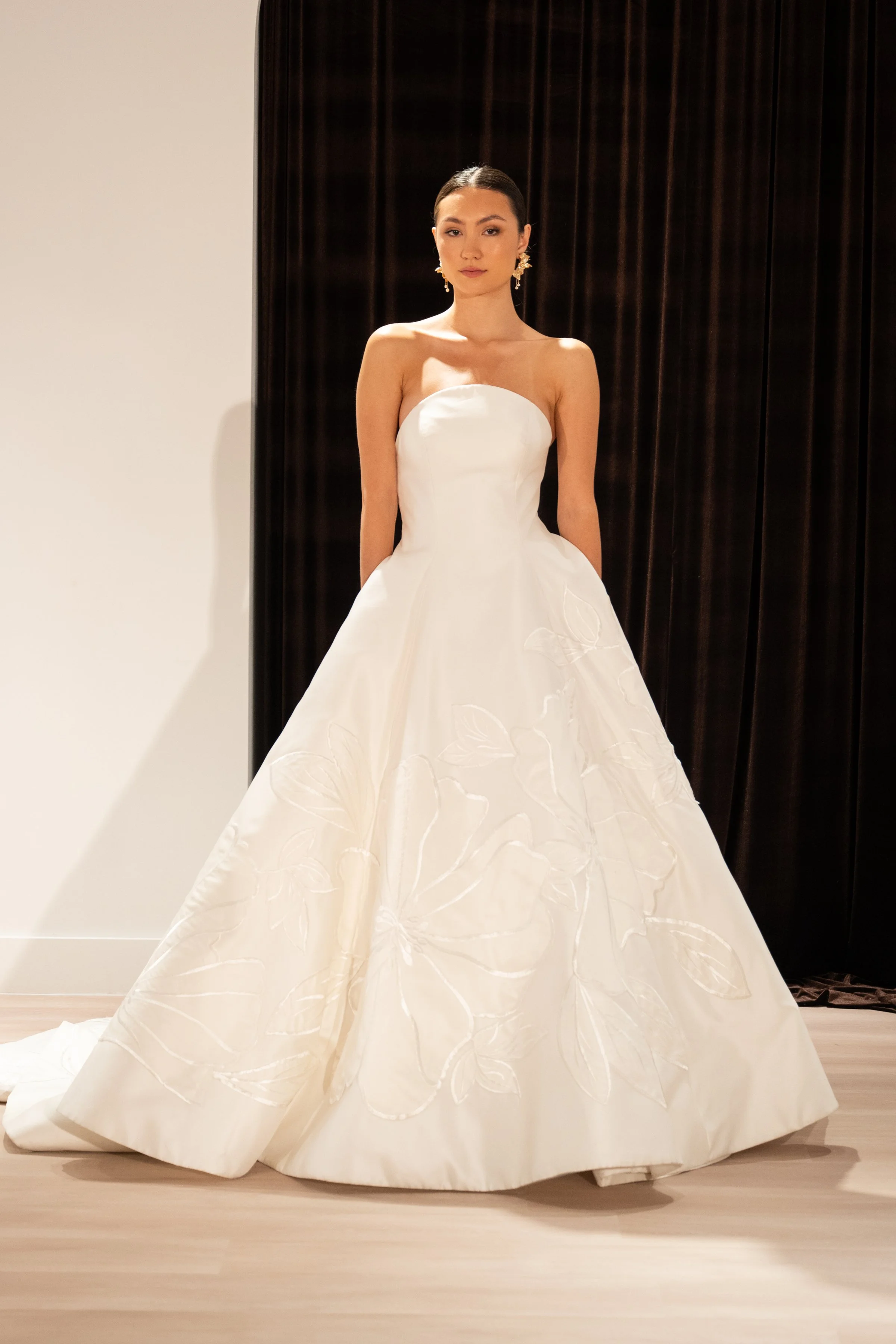 The Jenny Yoo Bridal Maya gown with applique is a modern yet timeless ball gown with a striking arched neckline and clean, elegant lines, anchored by a dramatic full skirt. Bold, oversized floral appliques artfully adorn the hem of the skirt, giving 
