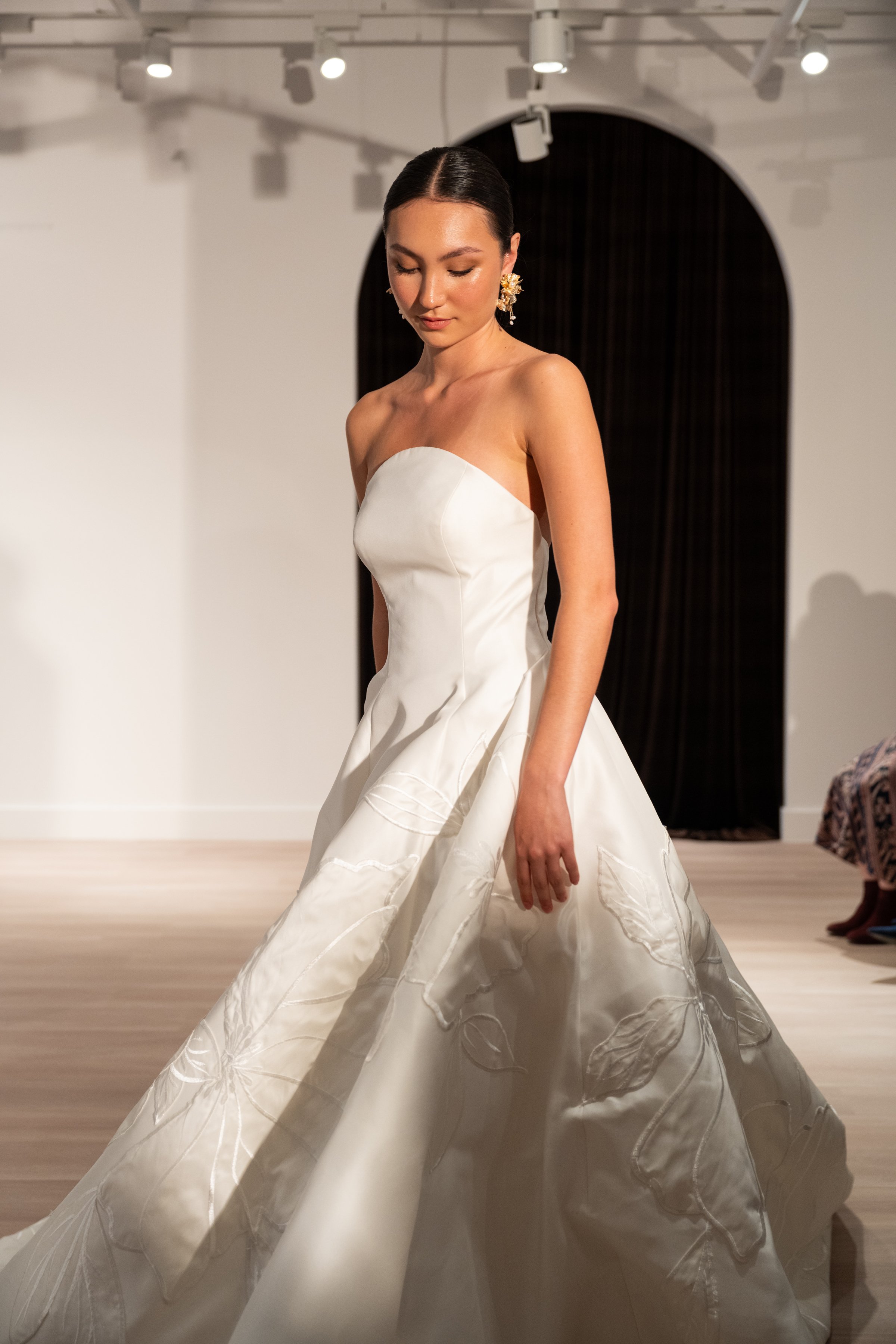The Jenny Yoo Bridal Maya gown with applique is a modern yet timeless ball gown with a striking arched neckline and clean, elegant lines, anchored by a dramatic full skirt. Bold, oversized floral appliques artfully adorn the hem of the skirt, giving 