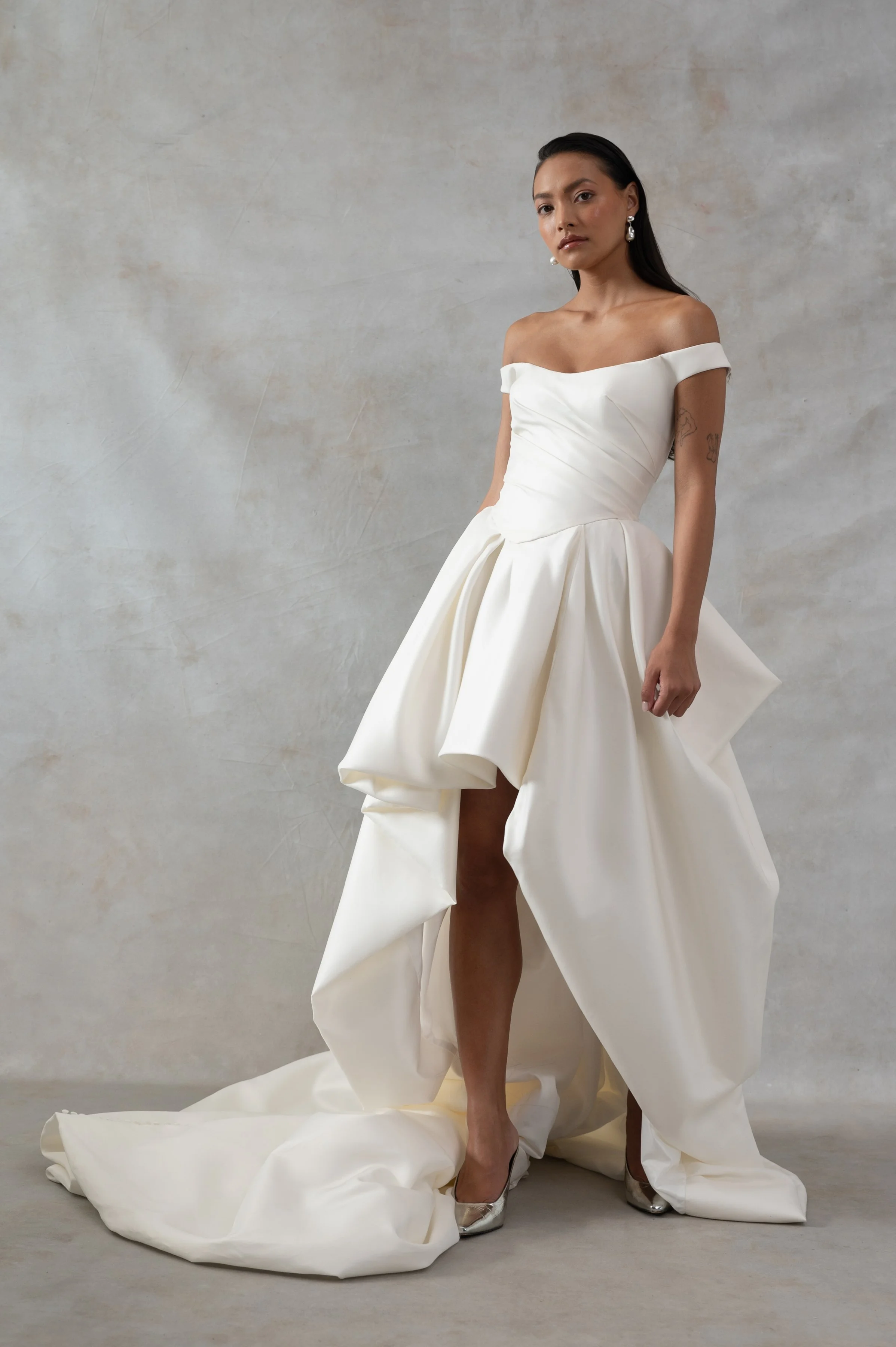 The Jenny Yoo Bridal Elle gown, cut in our lofty Italian taffeta, is a true chameleon. Featuring a corseted bodice with a flattering Basque waist and a delicate off the shoulder strap, her voluminous skirt transforms into a dramatic high-low look per
