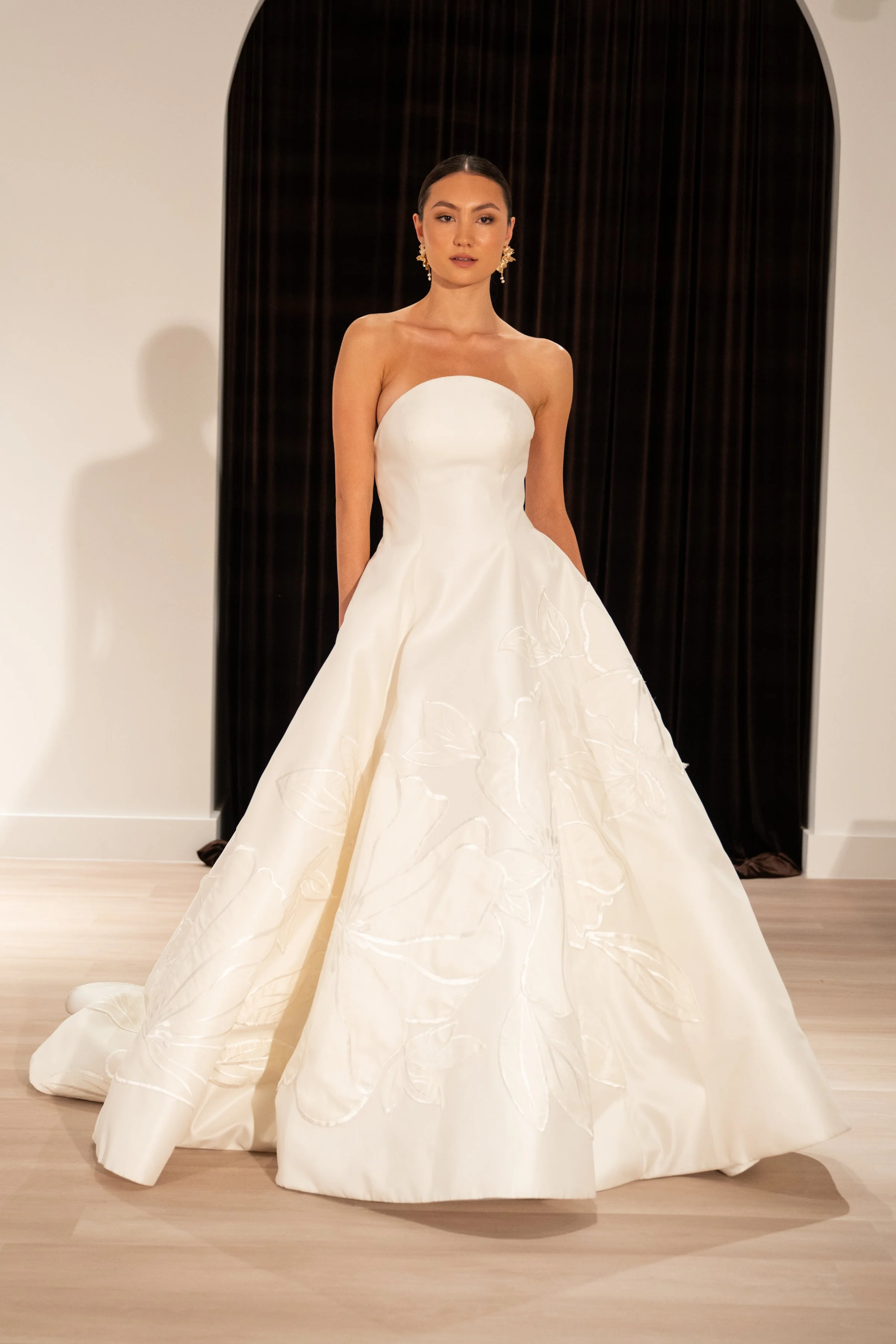 The Jenny Yoo Bridal Maya gown with applique is a modern yet timeless ball gown with a striking arched neckline and clean, elegant lines, anchored by a dramatic full skirt. Bold, oversized floral appliques artfully adorn the hem of the skirt, giving 
