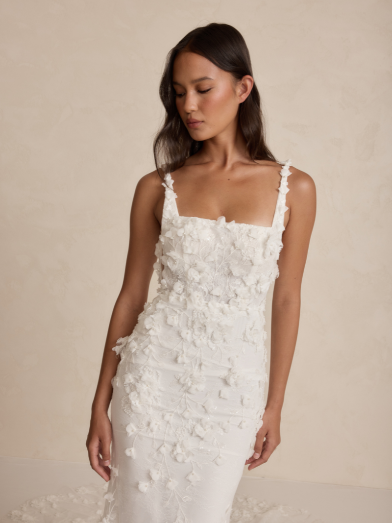 MWL Atlas features a bold square neckline and a signature low back with an illusion cut-out and delicate buttons. Crafted from unique 3D embroidered petal lace, it evokes nature's beauty. The fitted skirt enhances your curves, while the long train ad