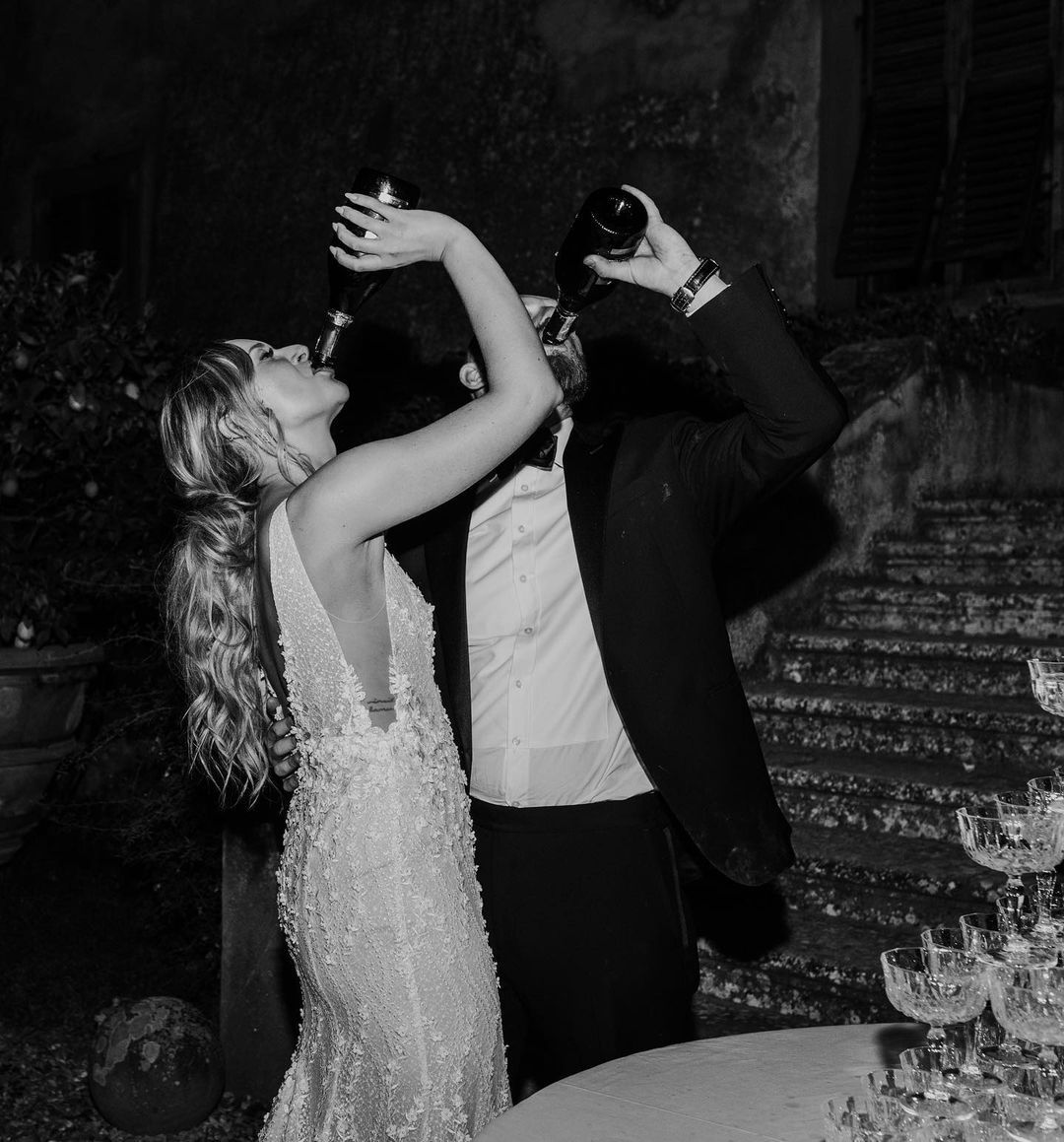 A man and a woman, likely at a formal event, are drinking from bottles. The woman is wearing a dress with lace details, and the man is dressed in a suit. They are standing outdoors near a staircase with potted plants and glassware on a table in the foreground.
