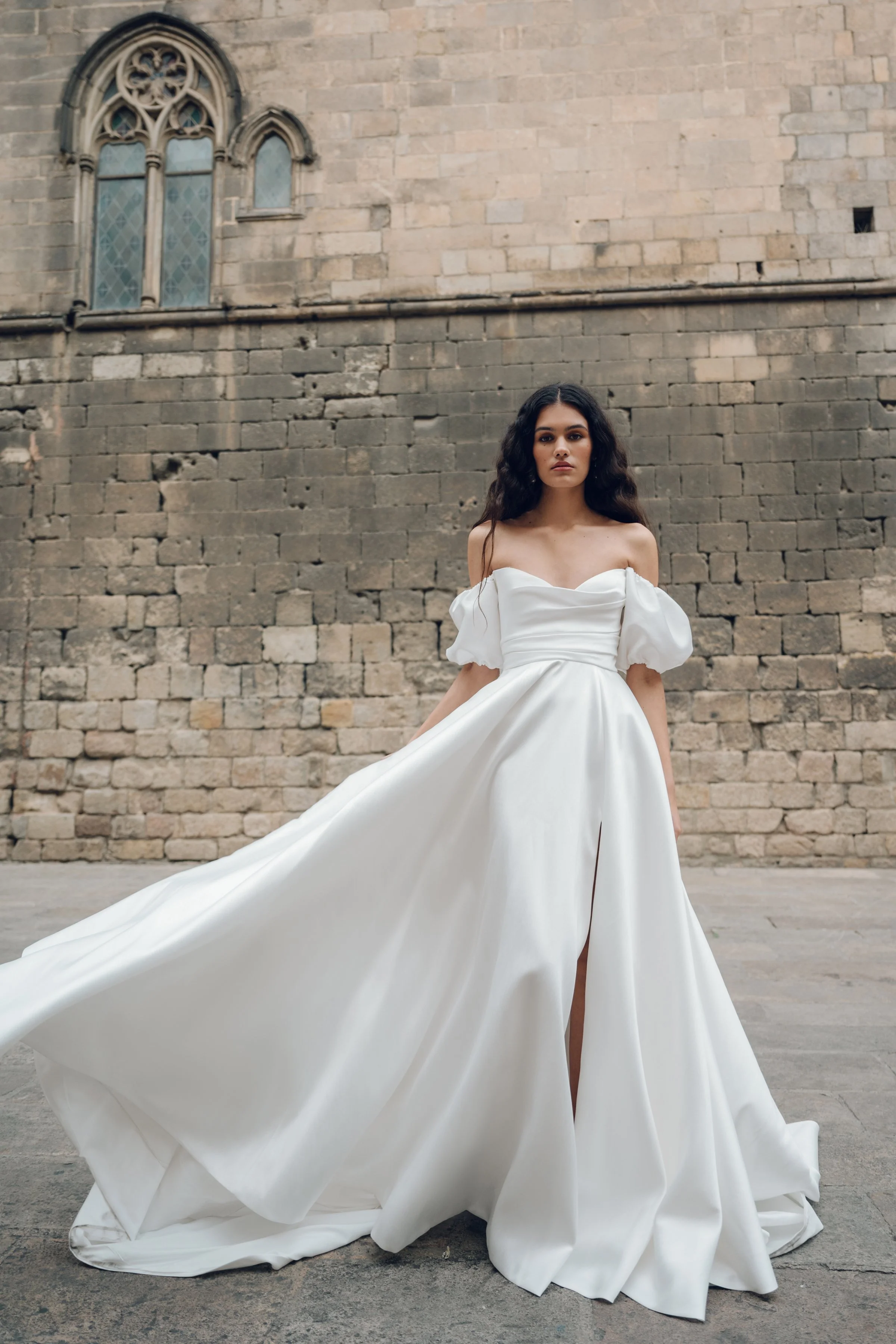 The Jenny Yoo Marisol gown is a romantic satin ball gown with a beautifully draped bodice and a sultry, sculpted cowl neckline that feels effortlessly feminine. It features a full skirt interrupted by a flattering slit and comes with detachable puff 