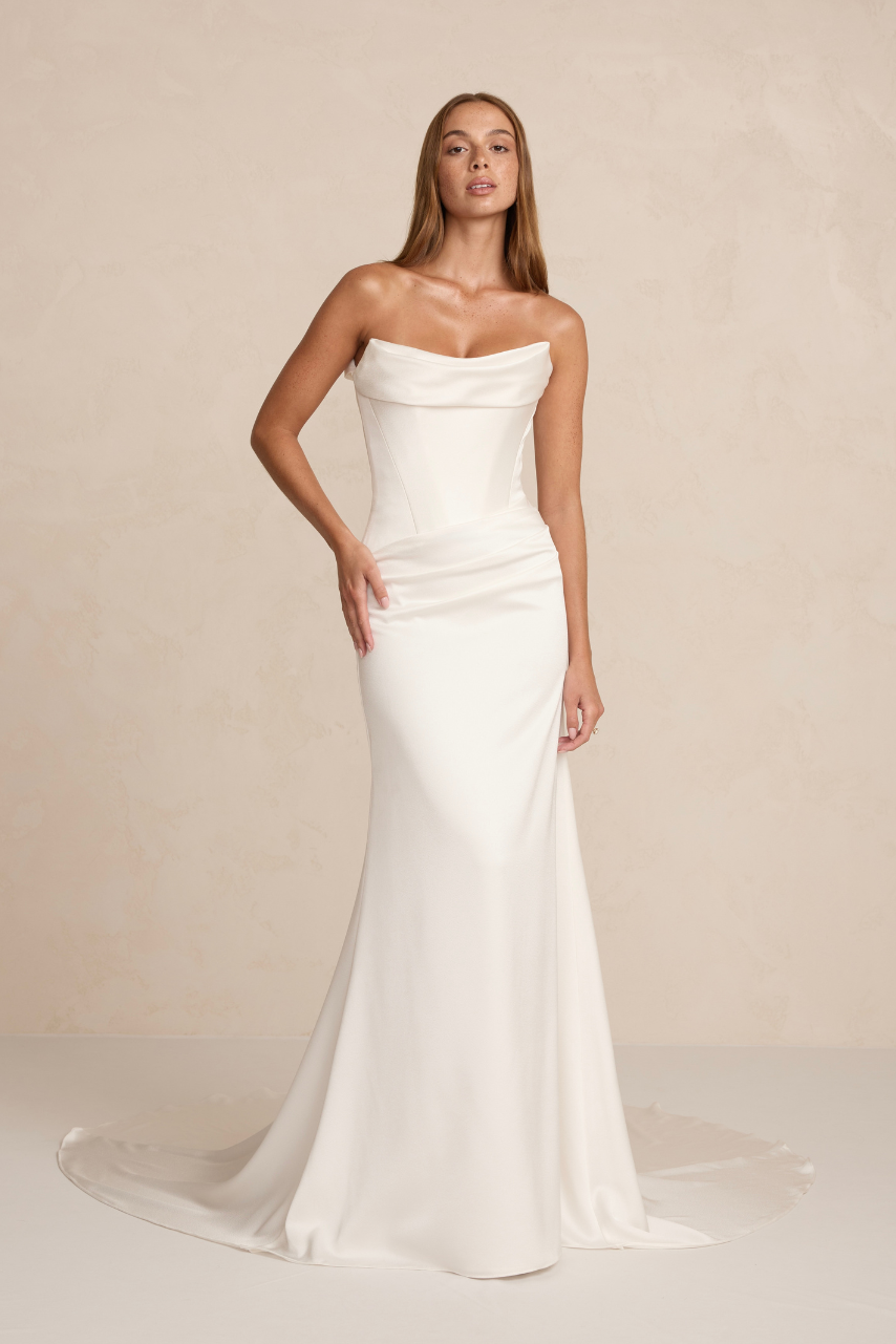The MWL Brooks is crafted from luxurious satin crepe, featuring an asymmetrical waistline, a structured boned bodice, and a soft front cowl. A sleek column skirt with four pleats creates an elegant, elongated silhouette. Available at Halo and Wren Br