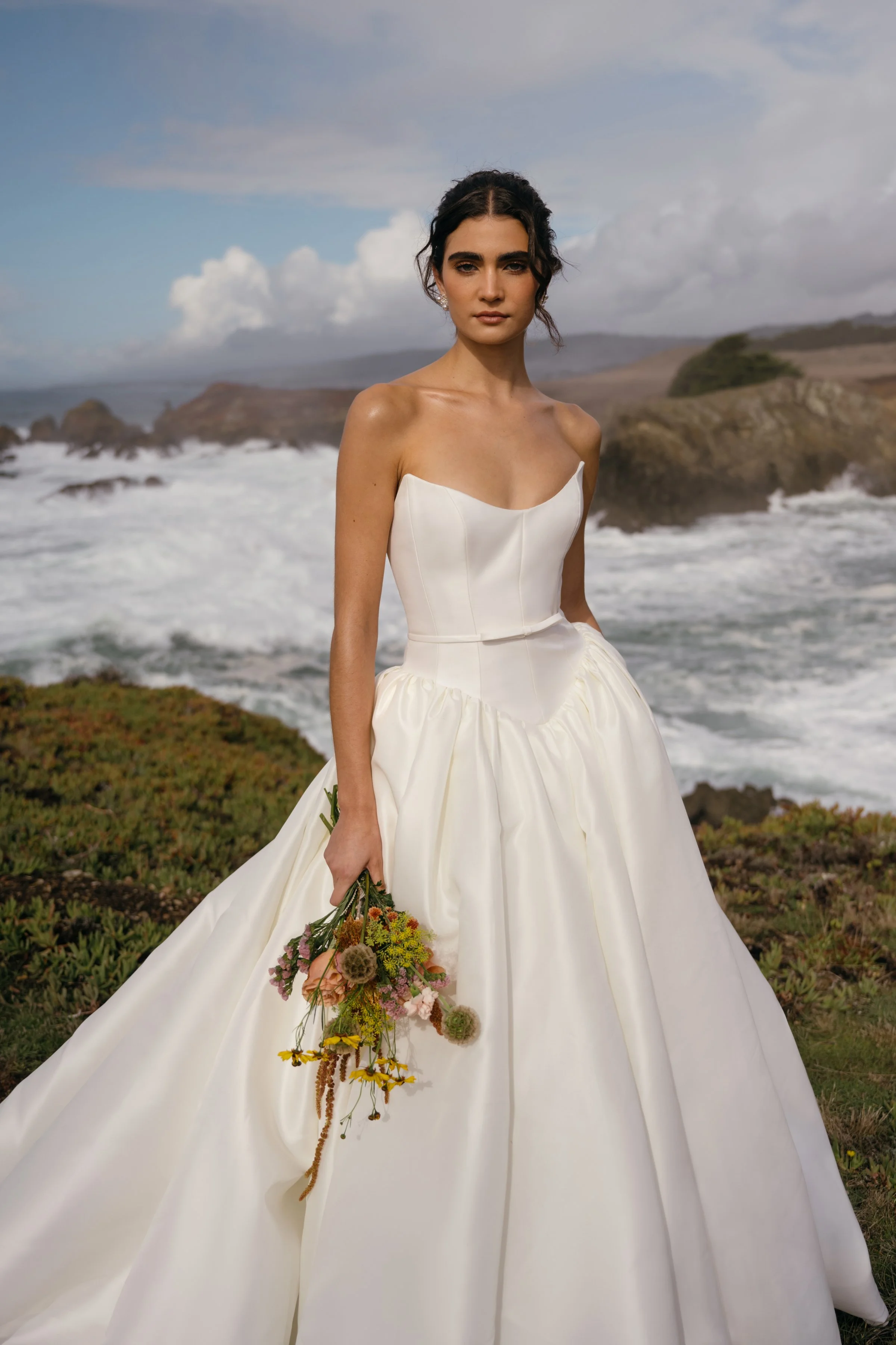 The Jenny Yoo Danielle gown is a romantic Italian taffeta ball gown with a fully corseted bodice and a striking high, peaked scoop neckline that creates a confident, flattering silhouette. It’s balanced by a voluminous gathered skirt, delicate bow sa