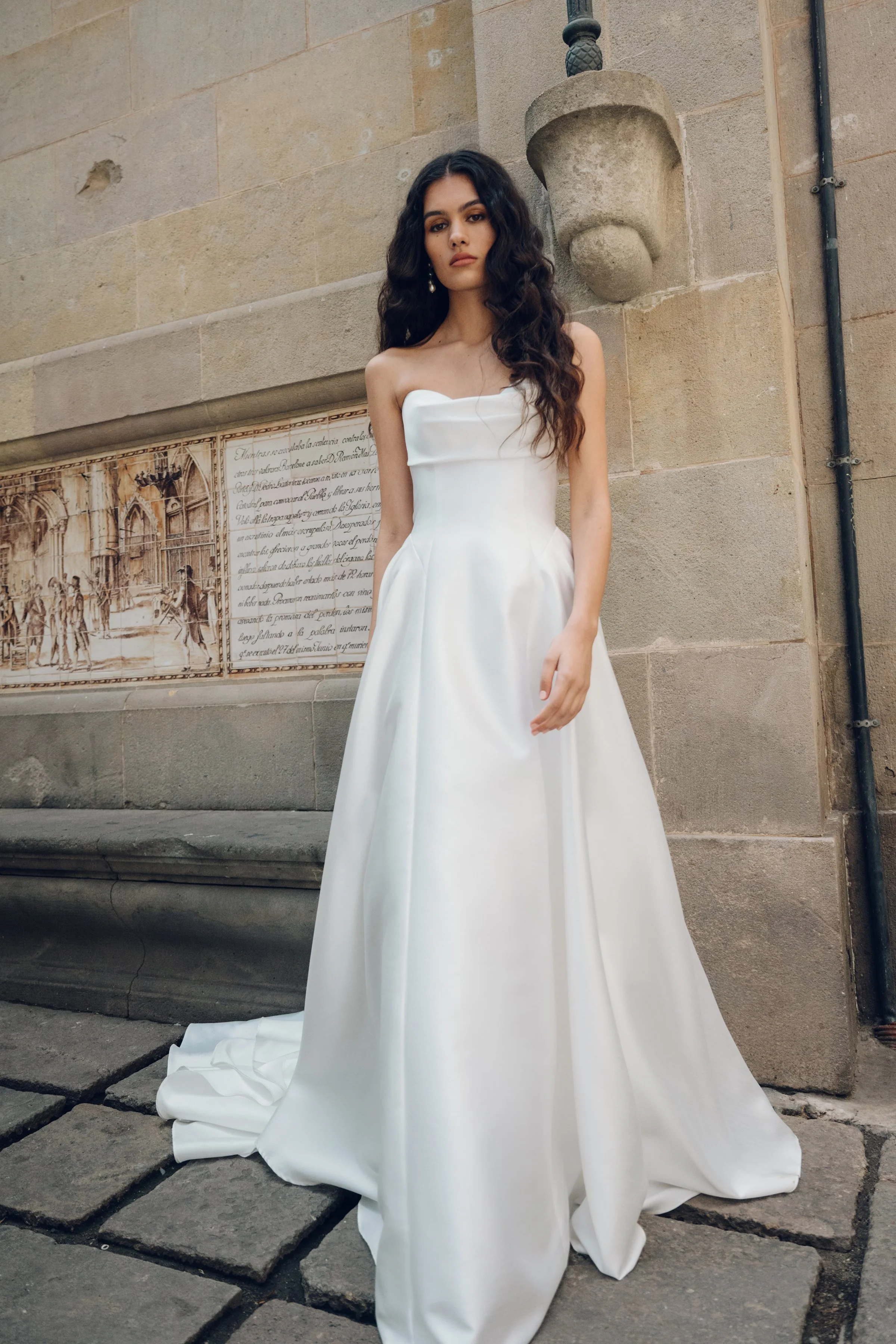 The Jenny Yoo Romina gown is a refined strapless A-line wedding dress crafted from lightweight, luminous canopy taffeta with a beautifully draped cowl bodice and sculpted drop-waist silhouette that feels both modern and timeless. Its soft, elegant li