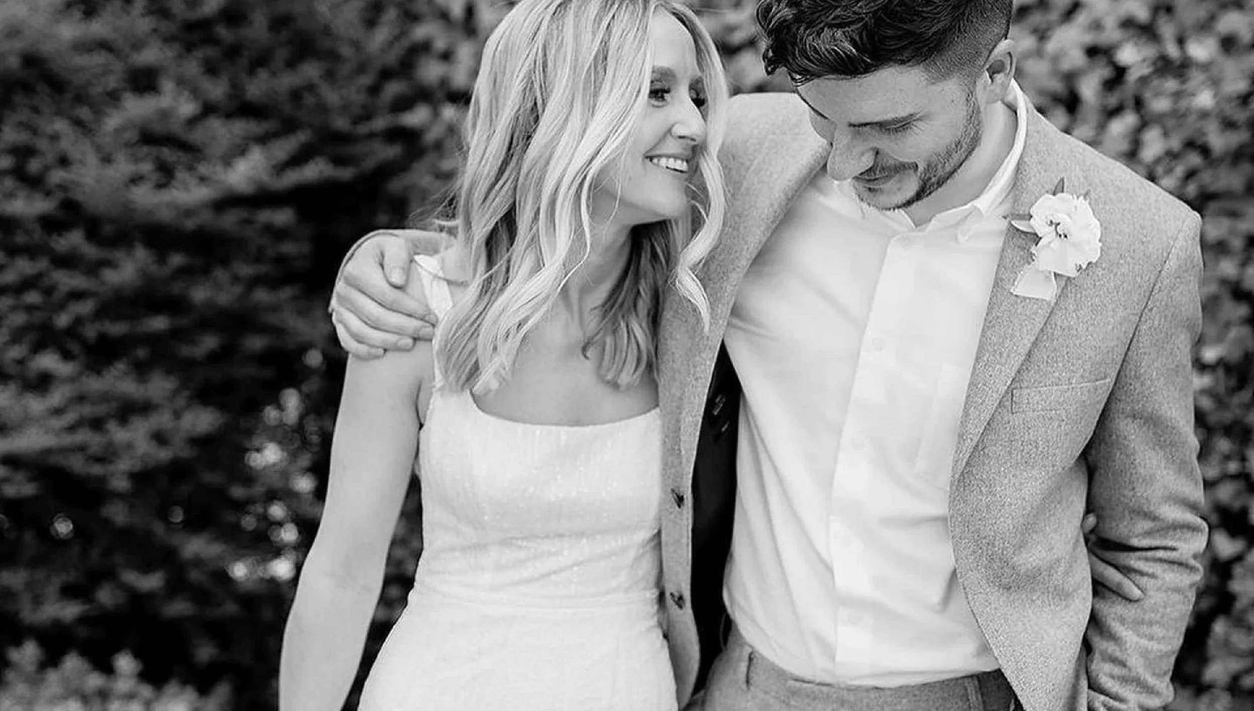 A black and white photo of a happy couple walking outdoors, with the woman wearing a sleeveless dress and the man in a suit jacket with a boutonniere, smiling and looking at each other.