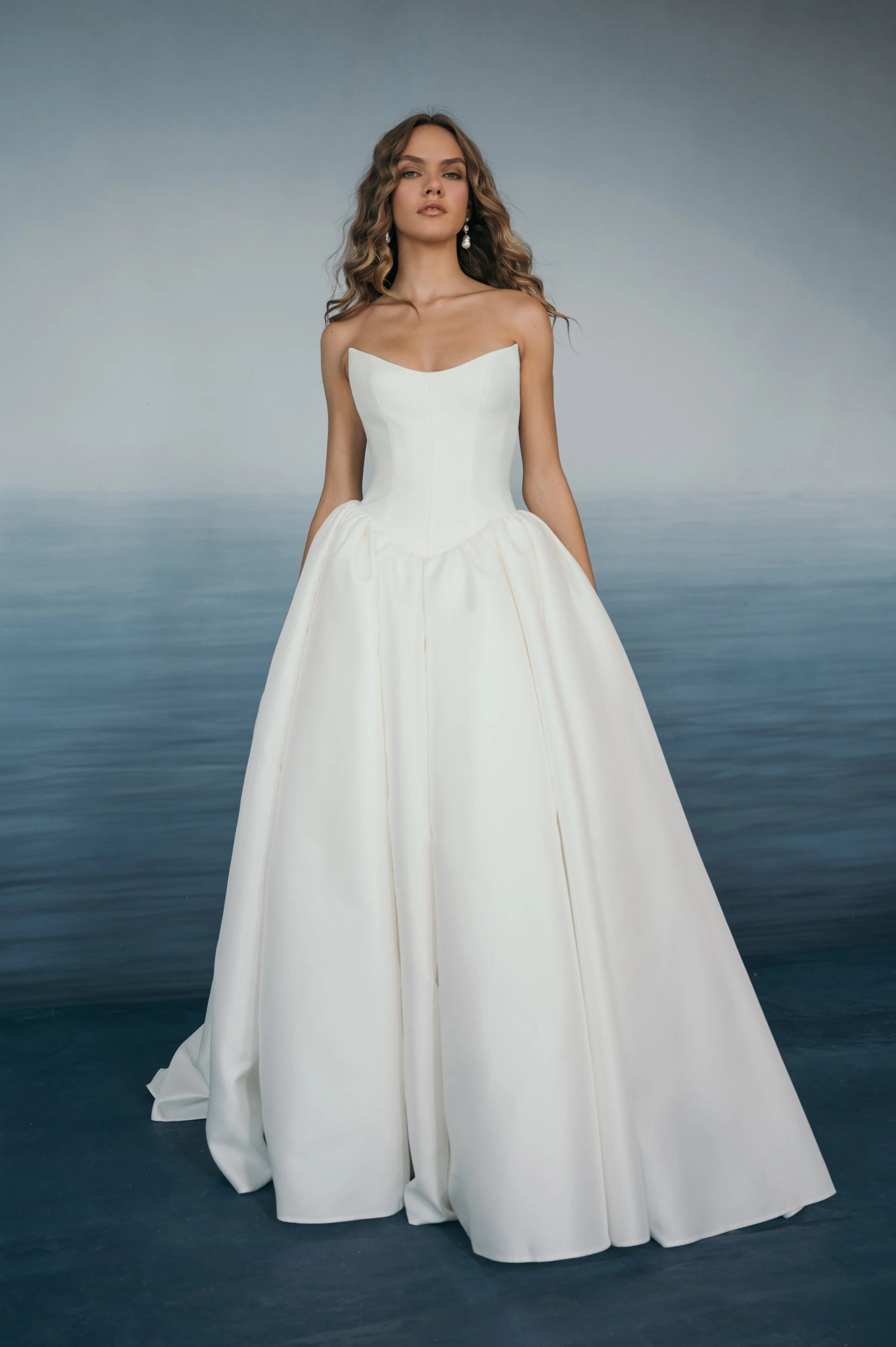 The Jenny Yoo Danielle gown is a romantic Italian taffeta ball gown with a fully corseted bodice and a striking high, peaked scoop neckline that creates a confident, flattering silhouette. It’s balanced by a voluminous gathered skirt, delicate bow sa