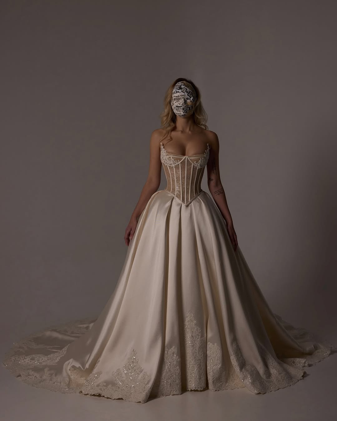 A woman in an elegant wedding gown with a corset-style bodice and full skirt, wearing a face mask with graffiti-style writing, standing against a neutral background.