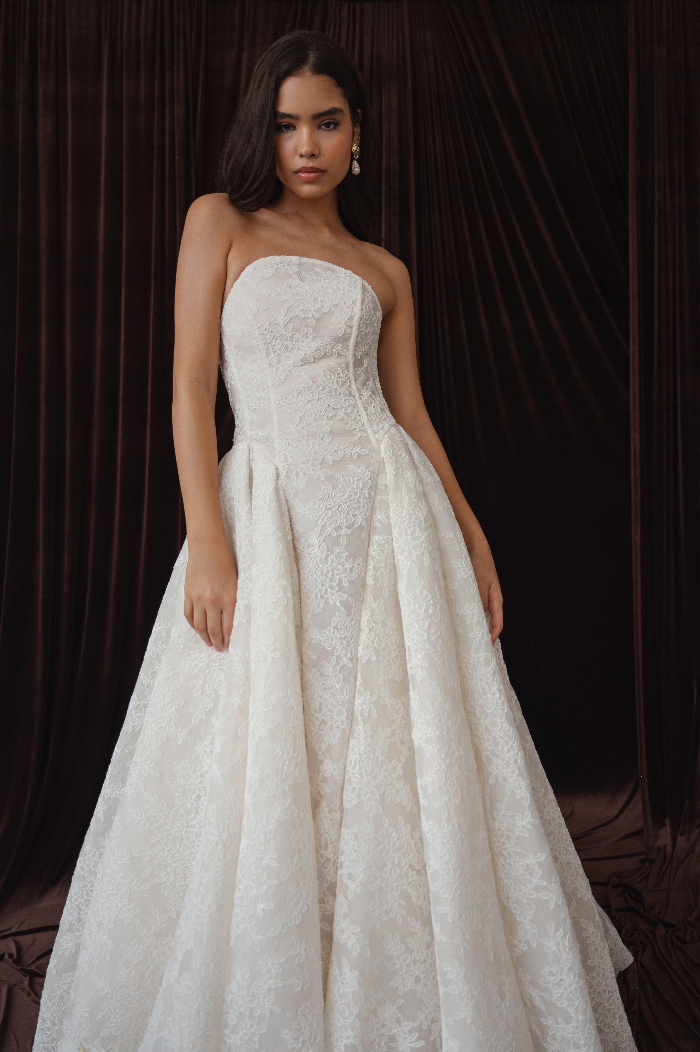 The Jenny Yoo Ember gown is a dramatic lace ball gown cut in intricately textured Chappell lace with a modern arched neckline and subtle drop-waist seaming that balances romantic tradition with contemporary flair. It features a full, pleated skirt wi