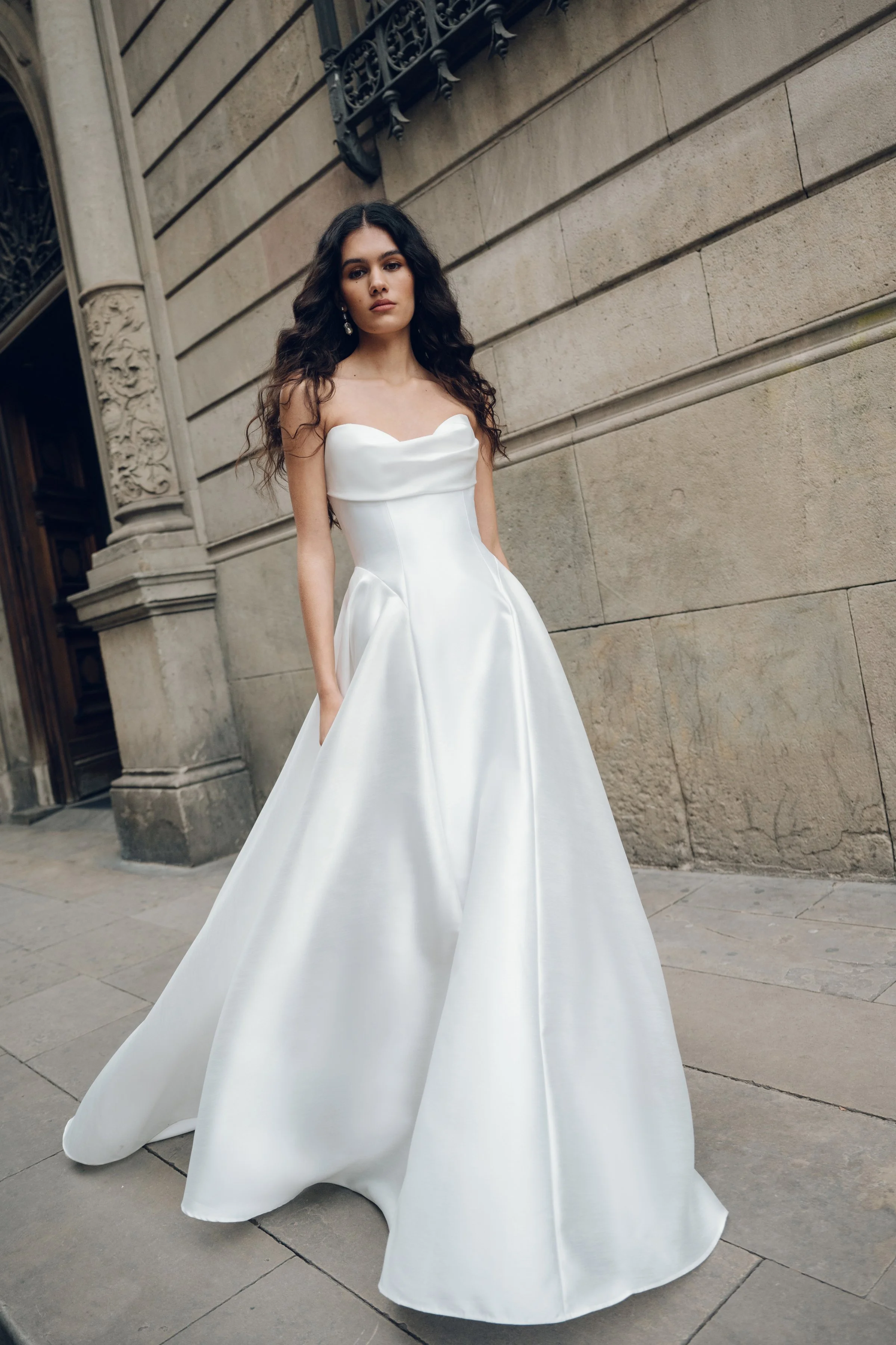 The Jenny Yoo Romina gown is a refined strapless A-line wedding dress crafted from lightweight, luminous canopy taffeta with a beautifully draped cowl bodice and sculpted drop-waist silhouette that feels both modern and timeless. Its soft, elegant li