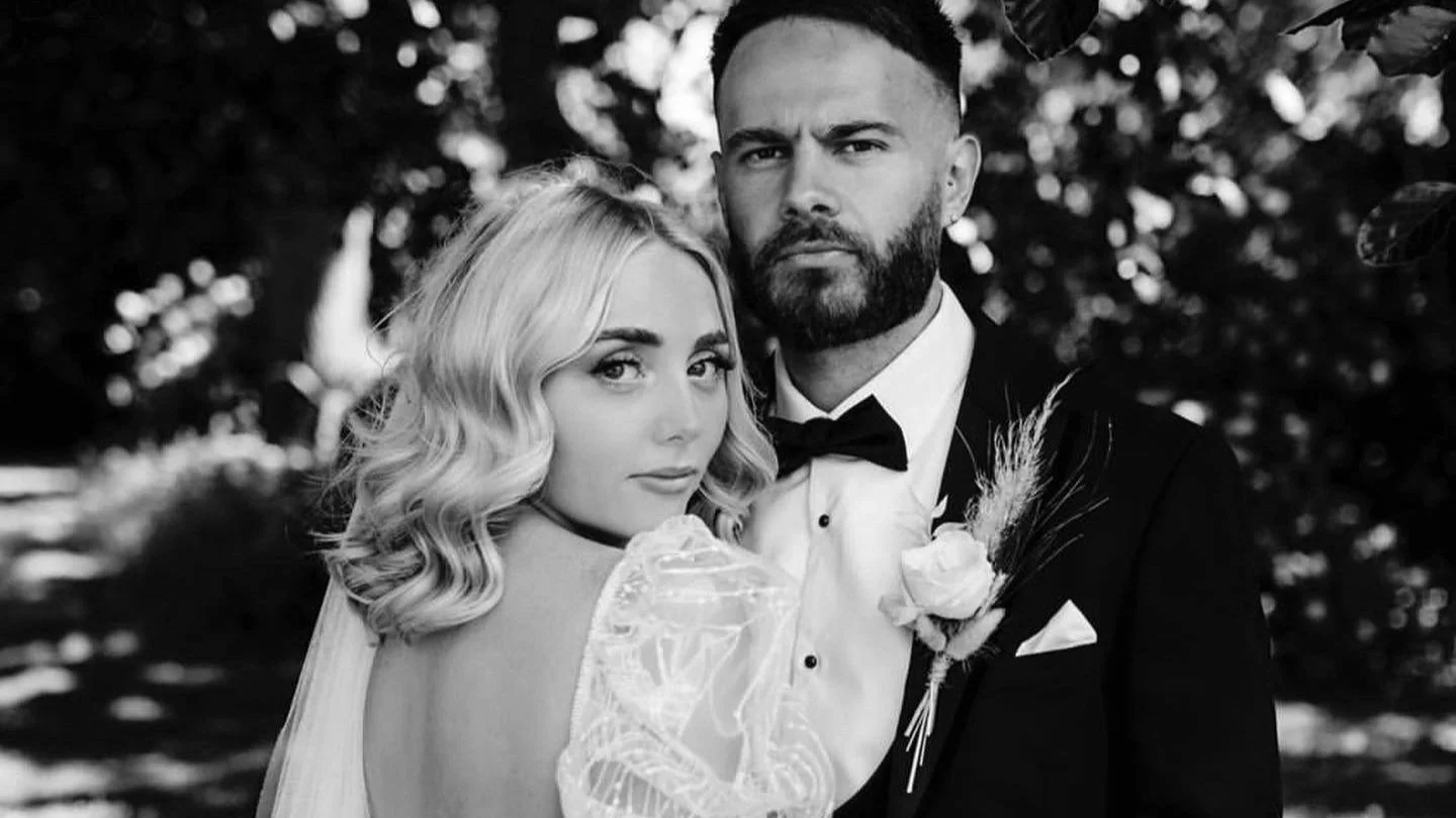 Black and white photo of a bride and groom in wedding attire, outdoors with trees in background, both looking at the camera.