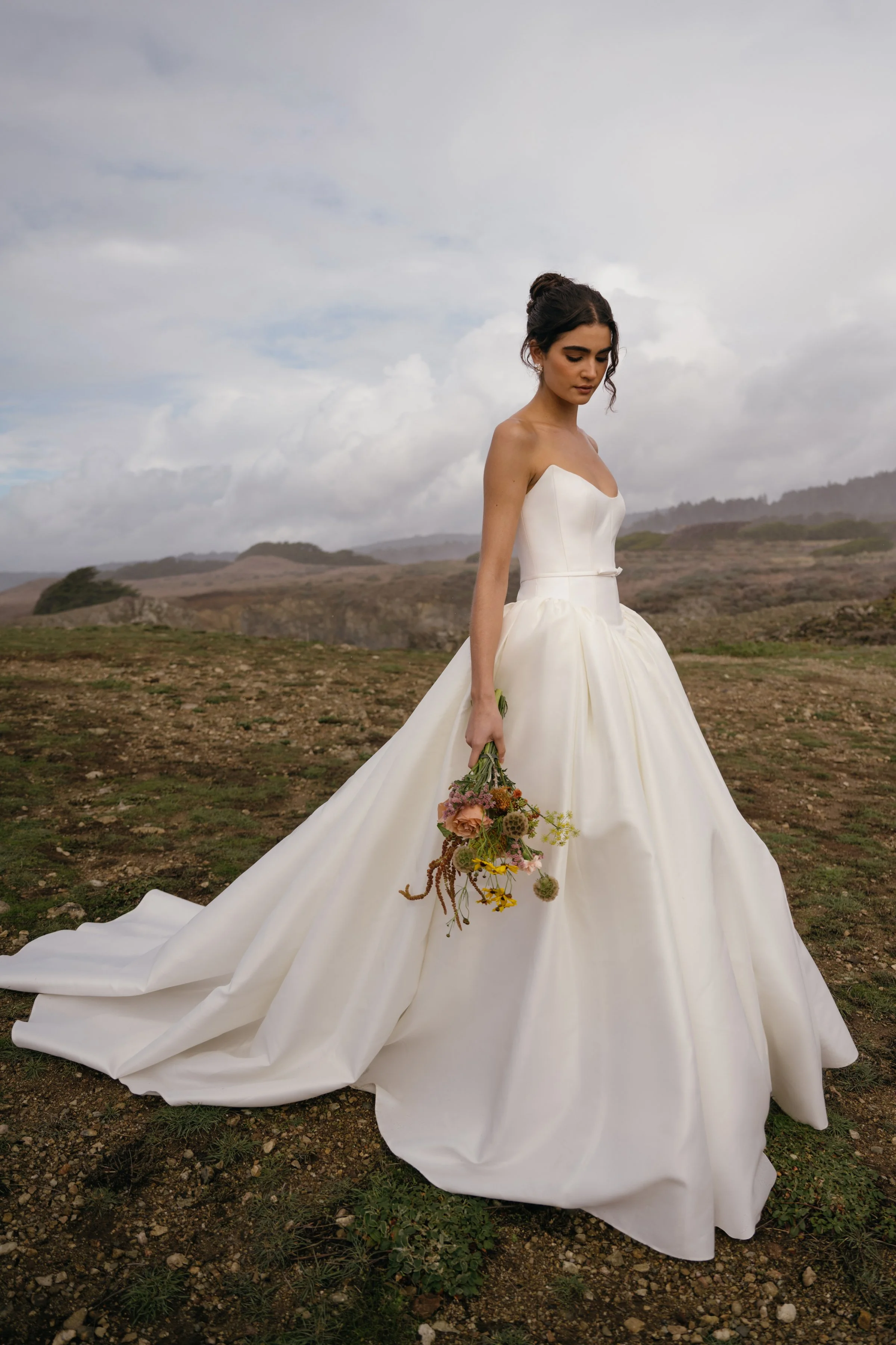 The Jenny Yoo Danielle gown is a romantic Italian taffeta ball gown with a fully corseted bodice and a striking high, peaked scoop neckline that creates a confident, flattering silhouette. It’s balanced by a voluminous gathered skirt, delicate bow sa