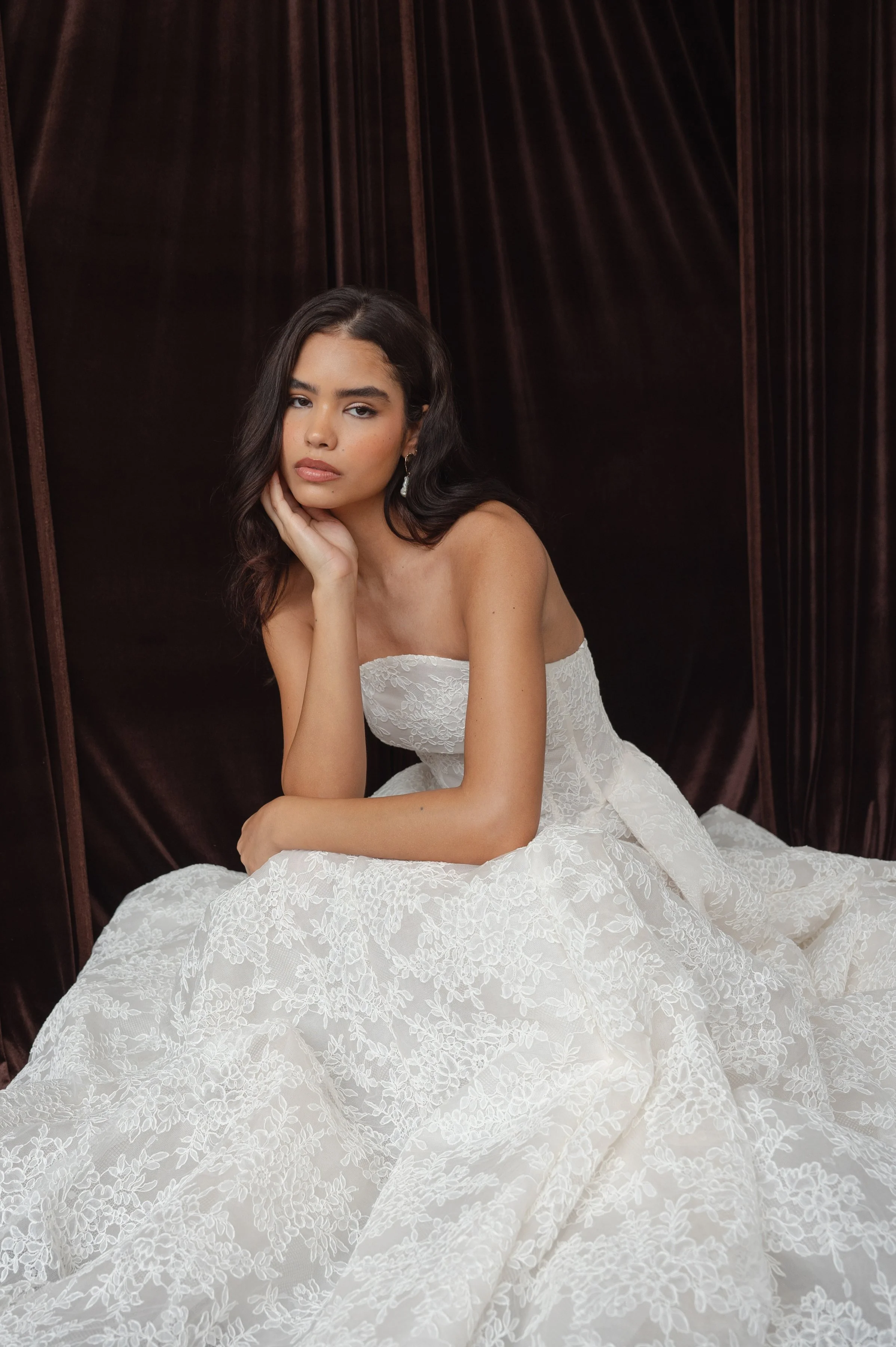 The Jenny Yoo Ember gown is a dramatic lace ball gown cut in intricately textured Chappell lace with a modern arched neckline and subtle drop-waist seaming that balances romantic tradition with contemporary flair. It features a full, pleated skirt wi