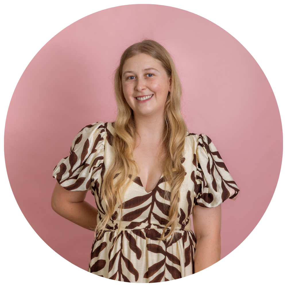 A smiling woman with long wavy blonde hair wearing a beige and brown patterned dress standing in front of a pink background.