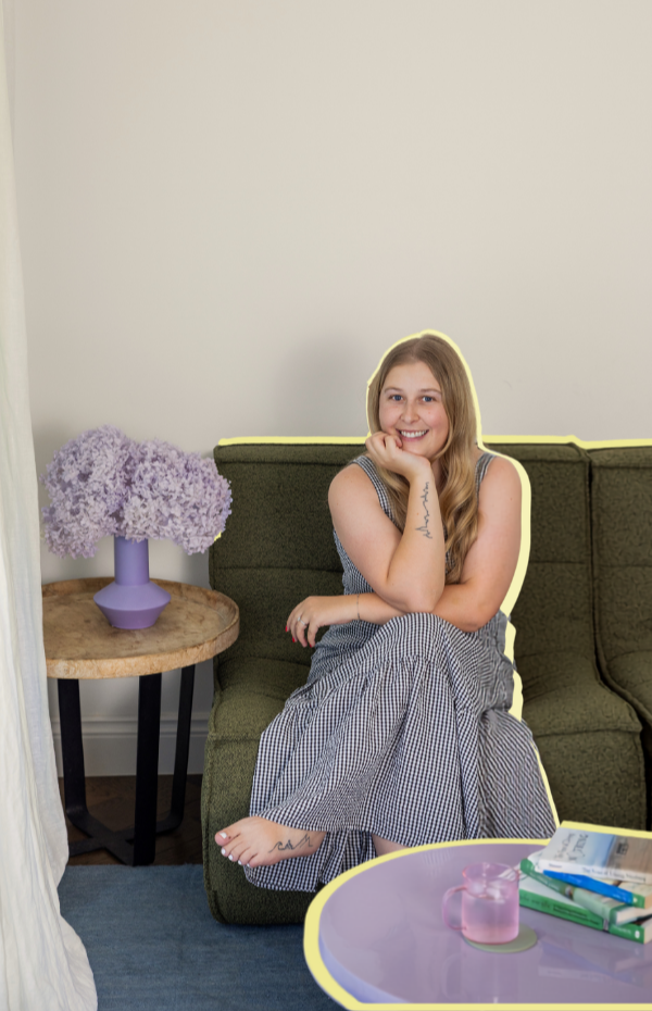 Smiling woman sitting on a green couch with her legs crossed, next to a side table with a purple vase holding purple flowers, and a purple coffee table with a pink mug and books.