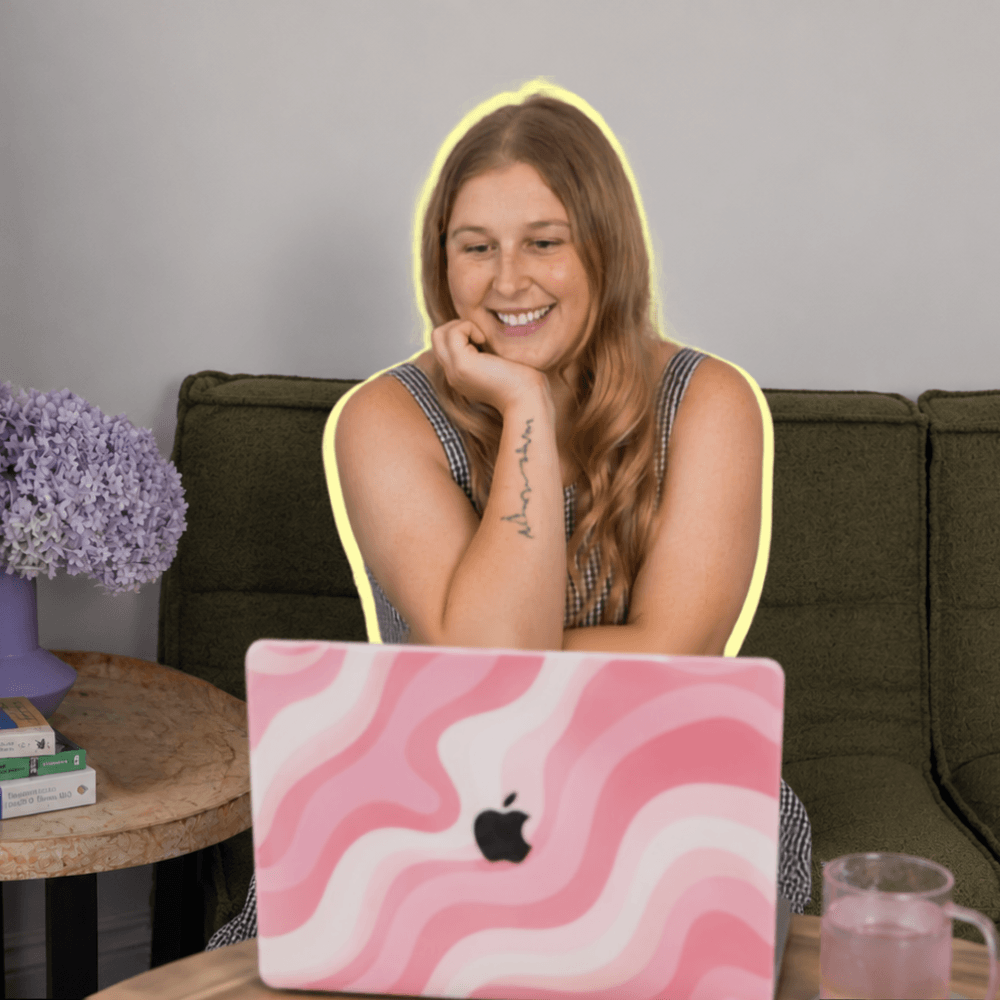 A woman with long red hair sitting on a green couch, smiling at her pink and white patterned MacBook, with purple flowers on a side table and a glass of water in front of her.