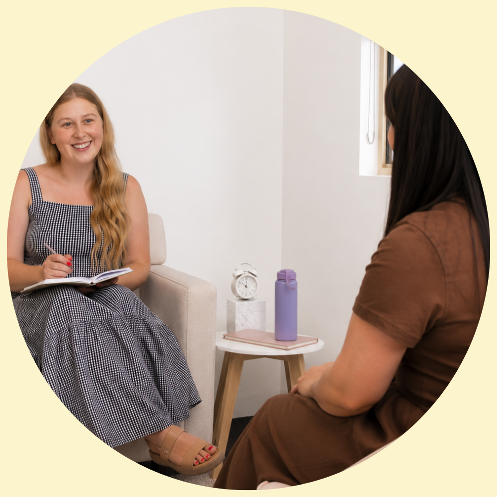 Two women having a conversation in a therapy or counseling session, one with long blonde hair in a checkered dress holding a notebook, and the other with darker straight hair in a brown shirt, sitting opposite each other in a bright room with a window, clock, and water bottle on a side table.