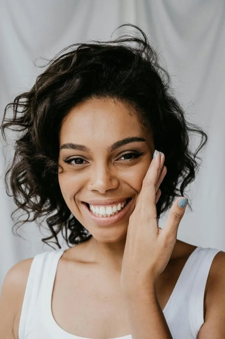 A woman with curly hair smiling and touching her face, wearing a white tank top.