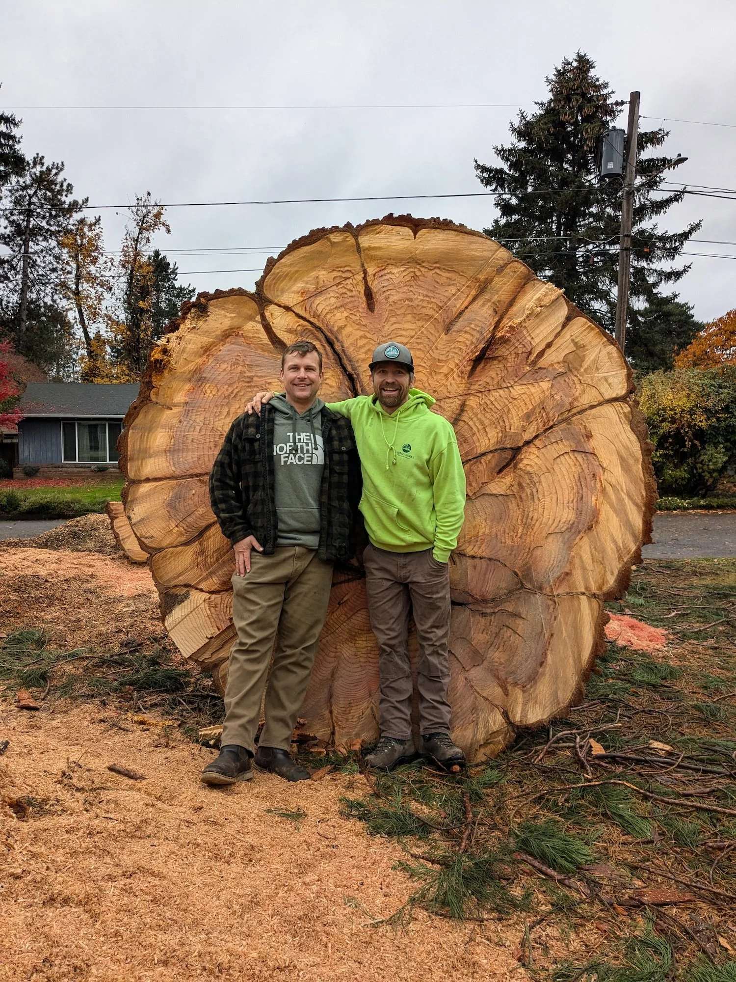 Two men standing in front of a large cut tree log, smiling at the camera. One is wearing a black plaid jacket and gray hoodie, and the other is wearing a bright green hoodie and gray cap.