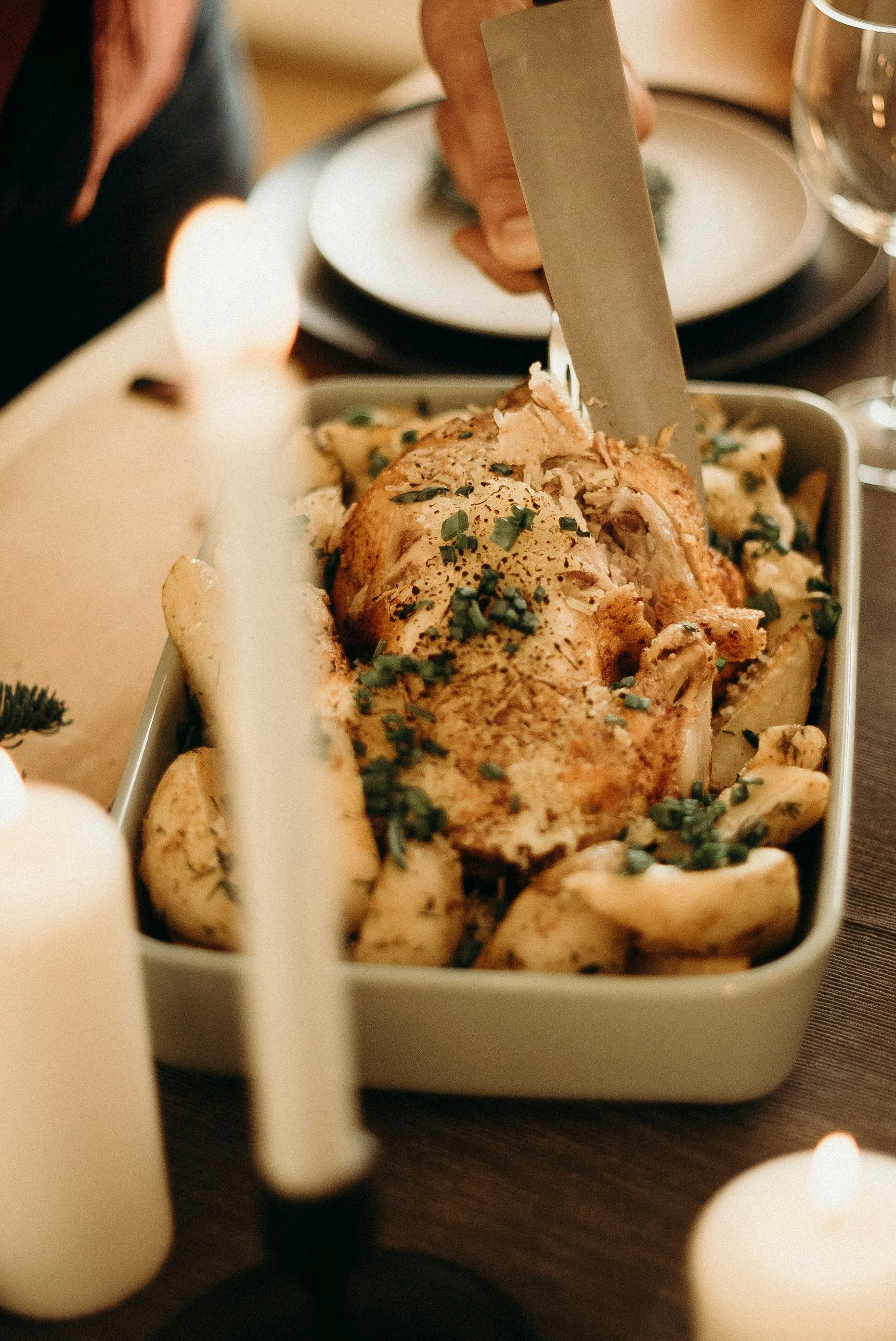 Person carving a roasted chicken with herbs in a baking dish, surrounded by lit candles and a dining setting.
