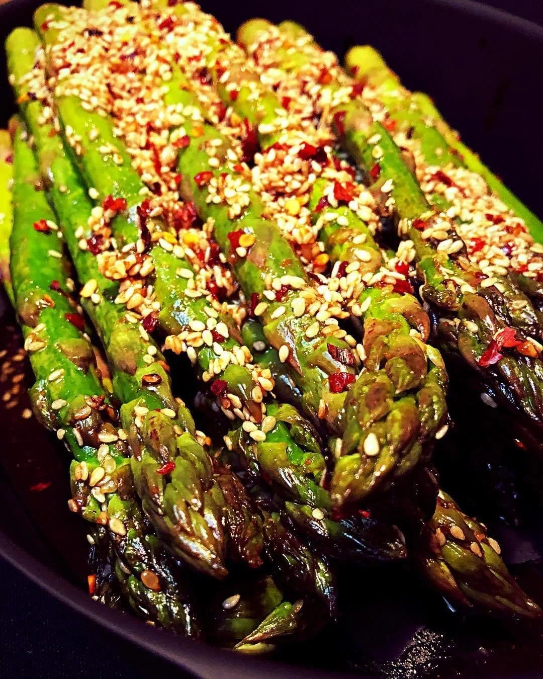 Roasted asparagus with sesame seeds and spices on a black plate.