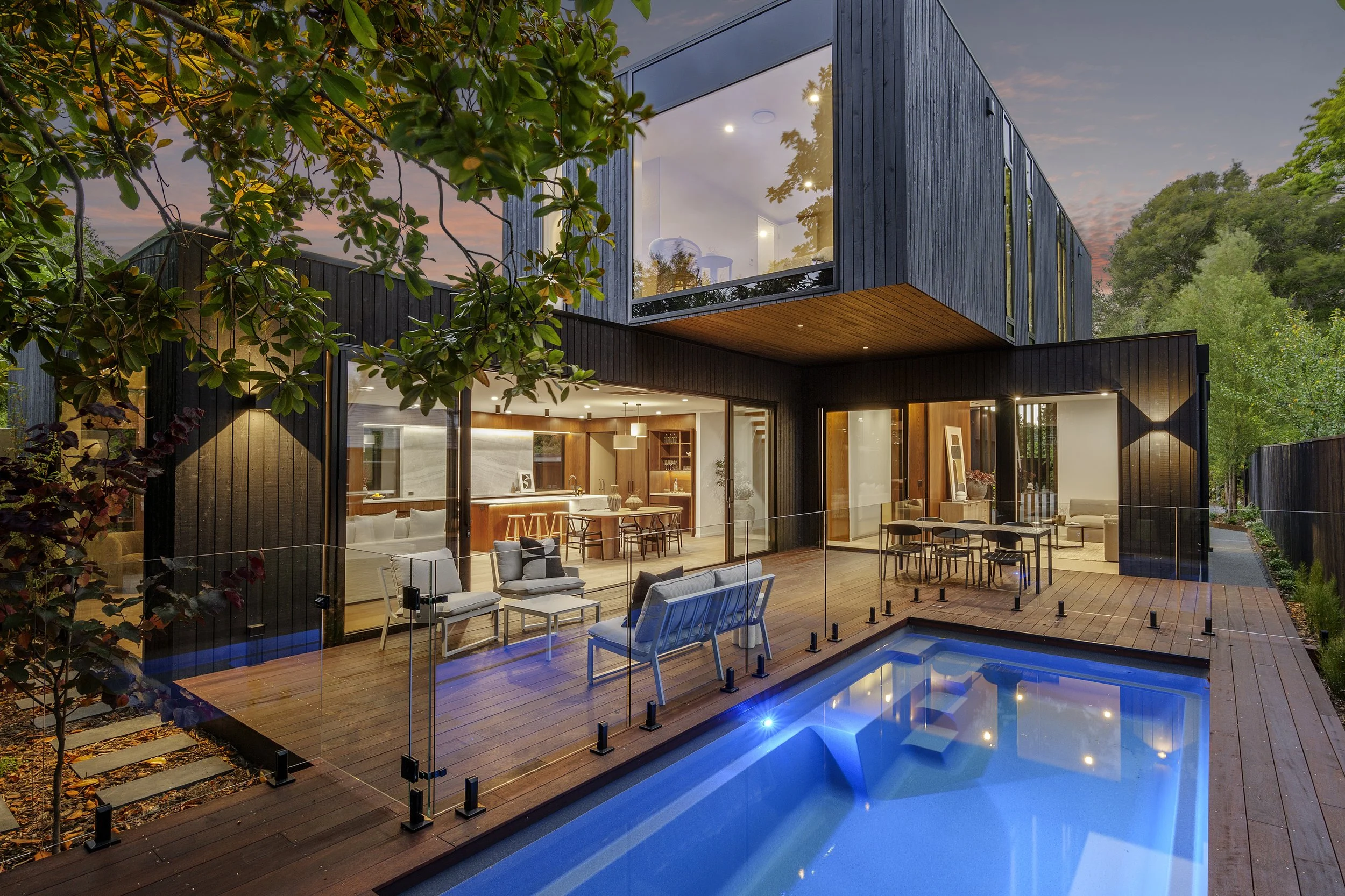 Modern two-story house with large windows, outdoor patio, and swimming pool, surrounded by trees, during sunset.
