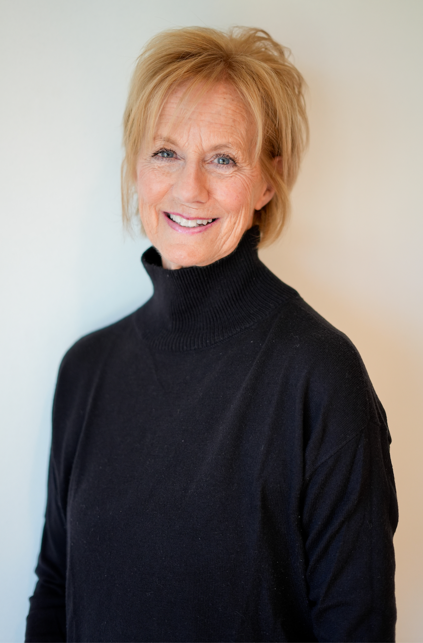 A smiling elderly woman with blonde hair in a black turtleneck sweater standing indoors next to a green houseplant.