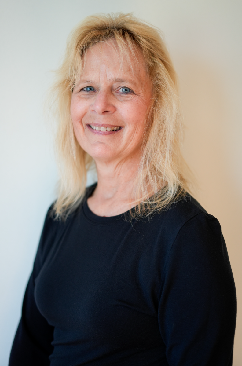 A woman with blonde hair and blue eyes standing outdoors in front of a concrete wall, smiling at the camera, wearing a black shirt and red plaid shirt.