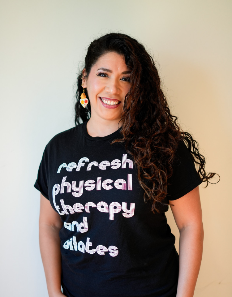 A woman with long, curly dark hair smiling and looking at the camera, wearing a sleeveless teal top and hoop earrings, standing in front of a plain gray background.
