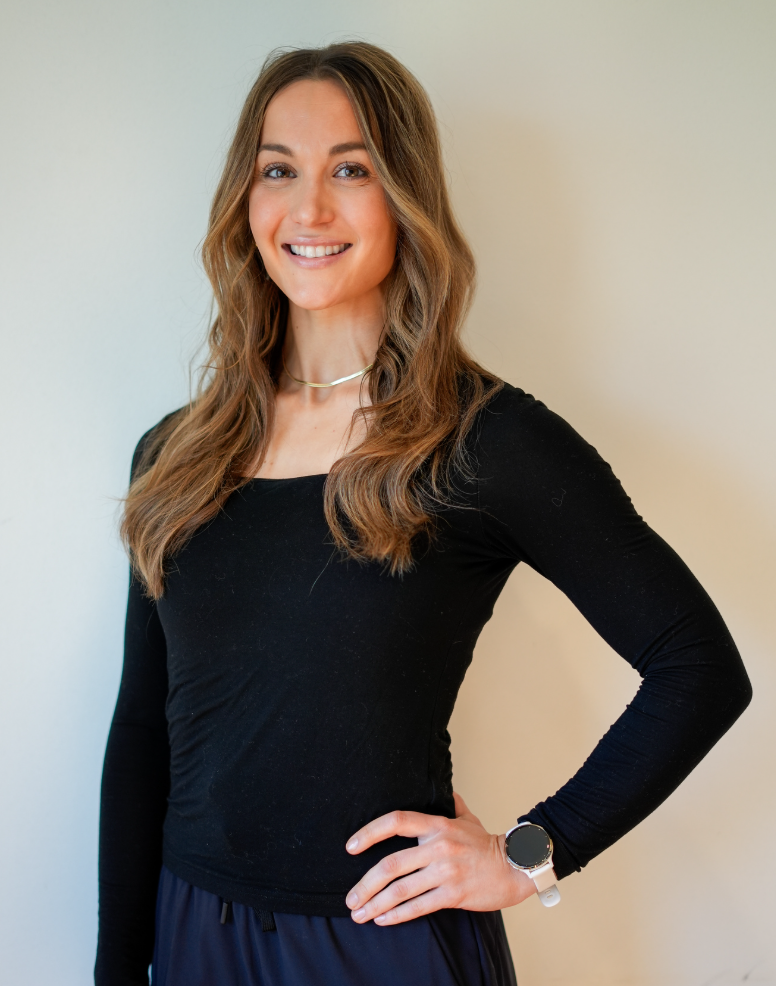 A smiling woman with long brown hair, wearing a white top, standing in front of a beige wall.
