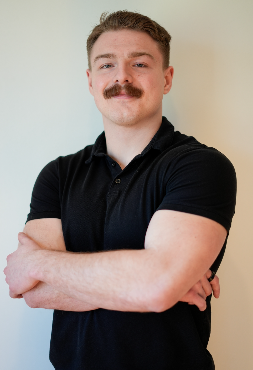 A man with a mustache and short hair stands against a textured white wall, crossing his arms and smiling at the camera, wearing a black T-shirt with white text.