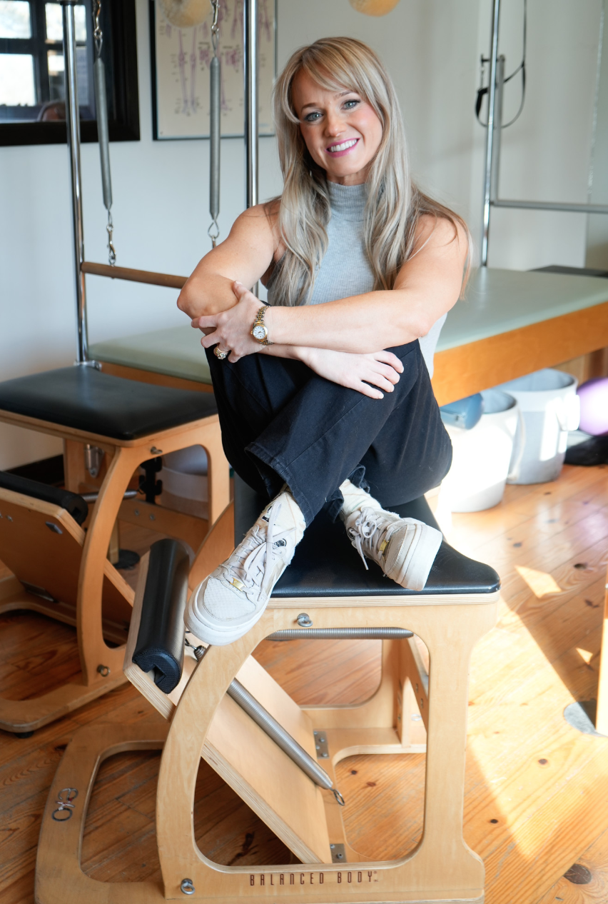 A woman with long blonde wavy hair smiling, sitting on a Pilates reformer machine, in a room with a window and light-colored walls.