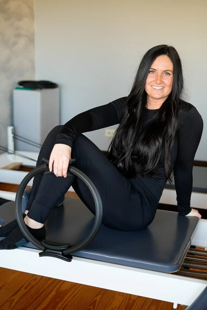 A woman with long brown hair and blue eyes, smiling, standing indoors next to a decorative brick wall and glass block window.